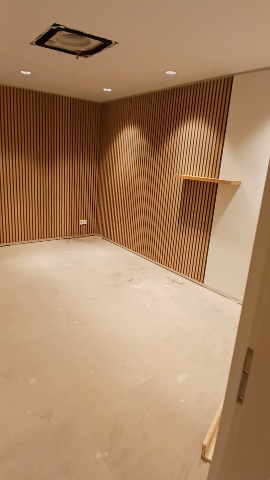 Pokój z wykończonymi ścianami z drewnianych paneli pionowych, z półką na jednej ze ścian. Podłoga w trakcie prac wykończeniowych, widoczne ślady po budowie. Na suficie widoczne oświetlenie punktowe...