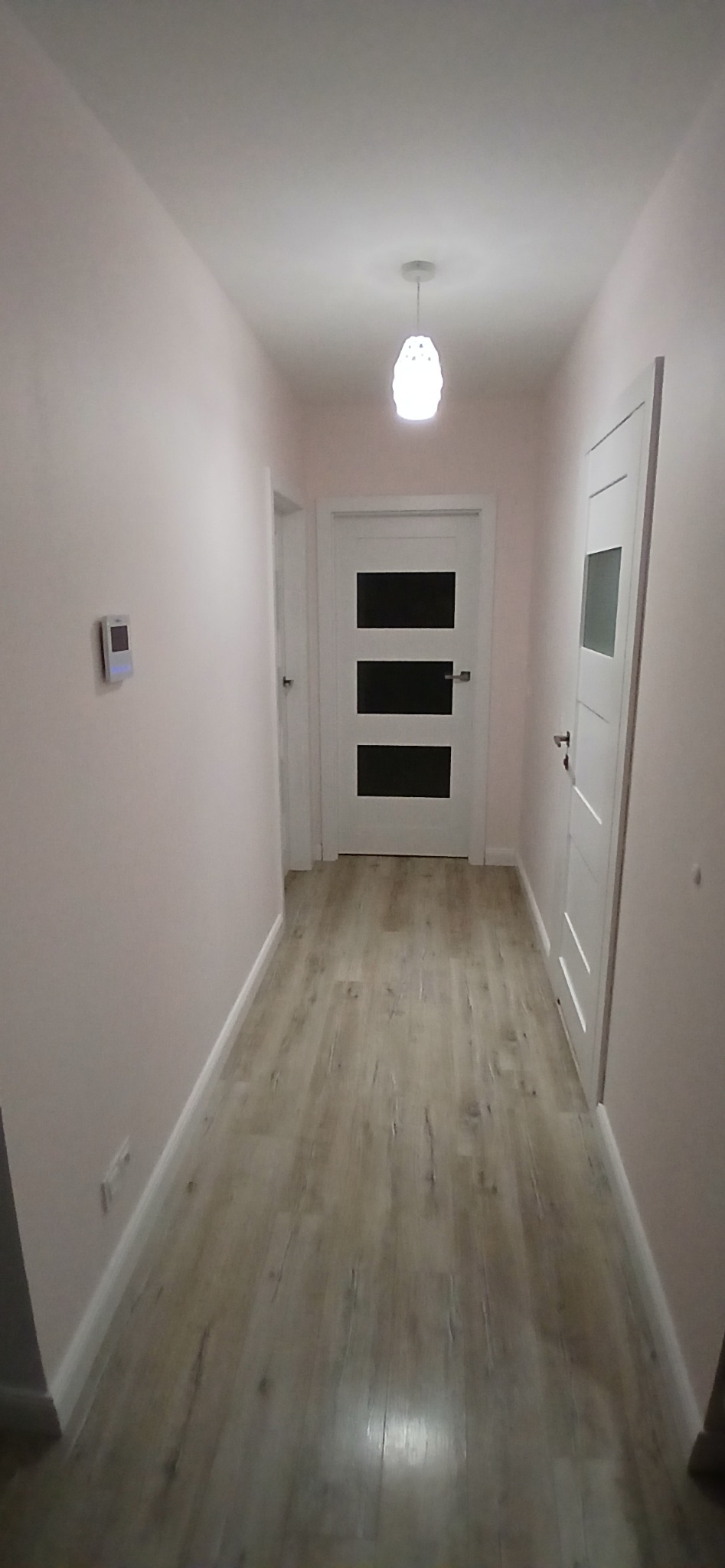 Korytarz po remoncie: świeżo pomalowane ściany w odcieniach różu, jasne panele podłogowe imitujące drewno, białe drzwi wewnętrzne z czarnymi wstawkami i wisząca lampa z ażurowym kloszem.