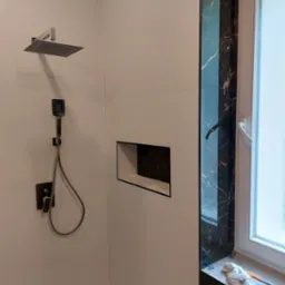 Nowoczesna łazienka z białymi płytkami, czarnymi akcentami marmuru wokół okna, wnęką w ścianie wykończoną czarnym marmurem oraz zestawem prysznicowym w kolorze ciemnego grafitu.