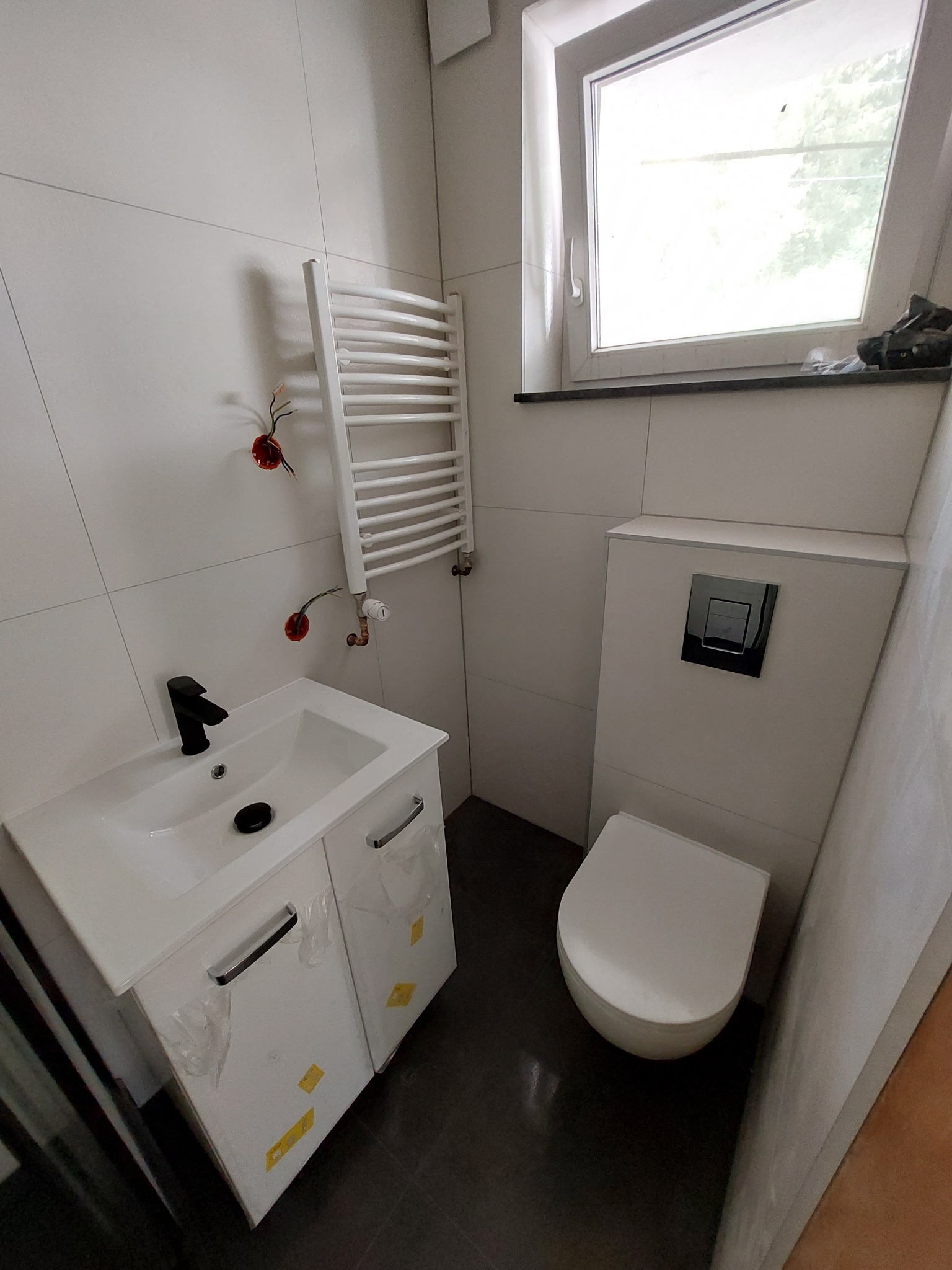 Nowoczesna łazienka z białymi płytkami, wiszącą toaletą z czarnym przyciskiem spłukiwania, umywalką z czarną baterią i grzejnikiem drabinkowym; widoczne niedokończone podłączenia elektryczne.