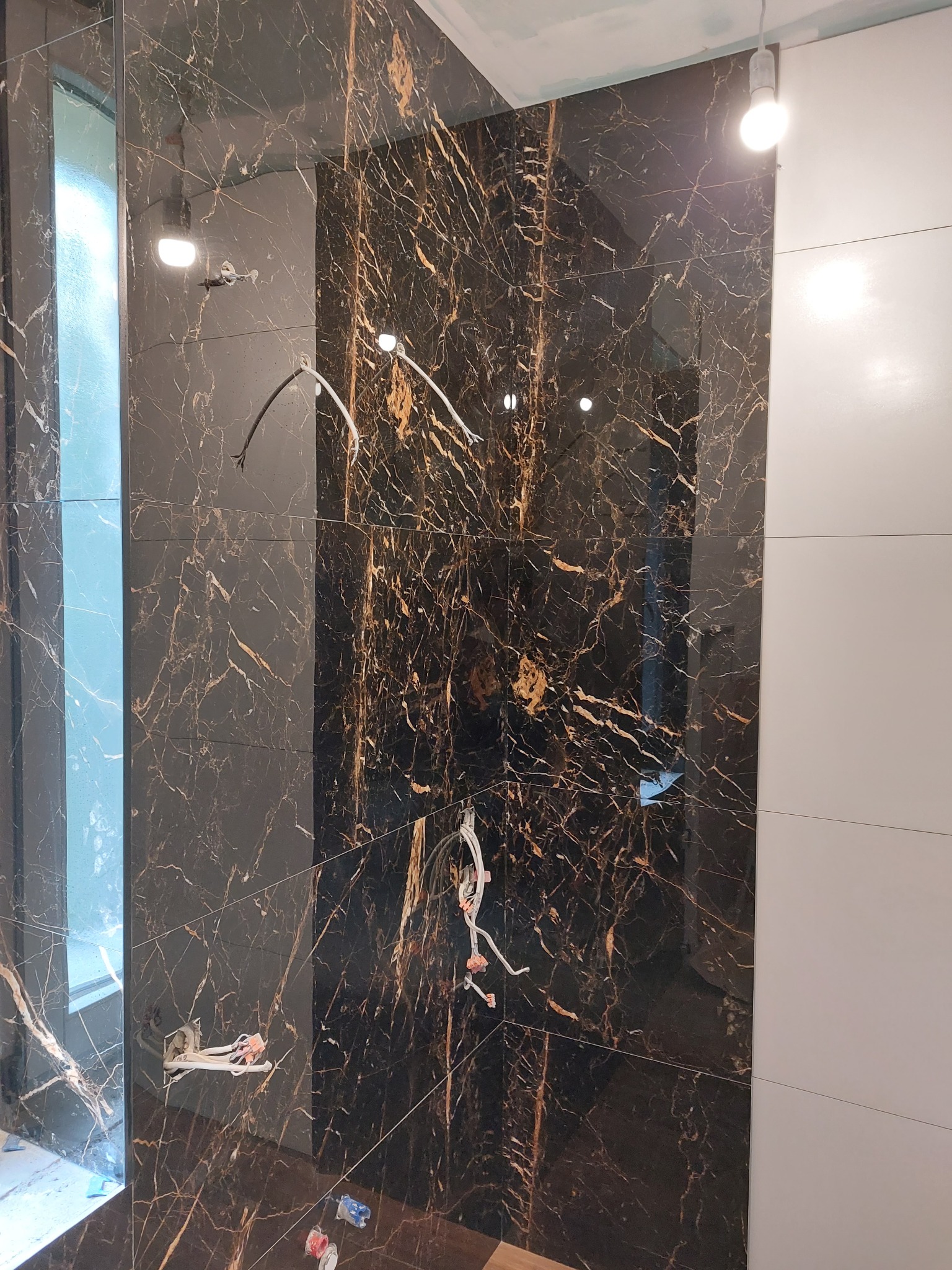 Ściana w trakcie układania płytek imitujących marmur z widocznymi kablami elektrycznymi i fragmentem białych płytek po prawej stronie.