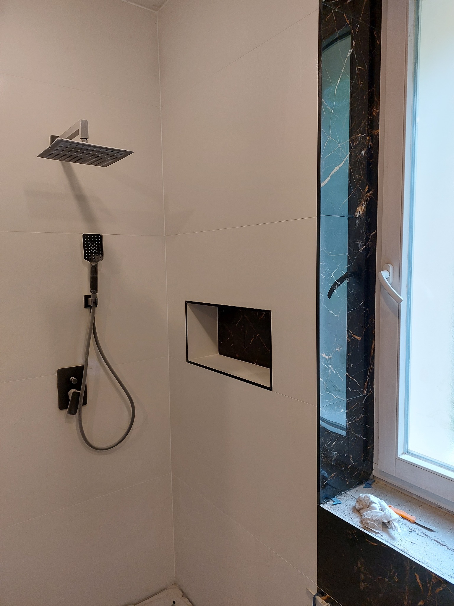 Nowoczesna łazienka z białymi płytkami, czarnymi akcentami marmuru wokół okna, wnęką w ścianie wykończoną czarnym marmurem oraz zestawem prysznicowym w kolorze ciemnego grafitu.