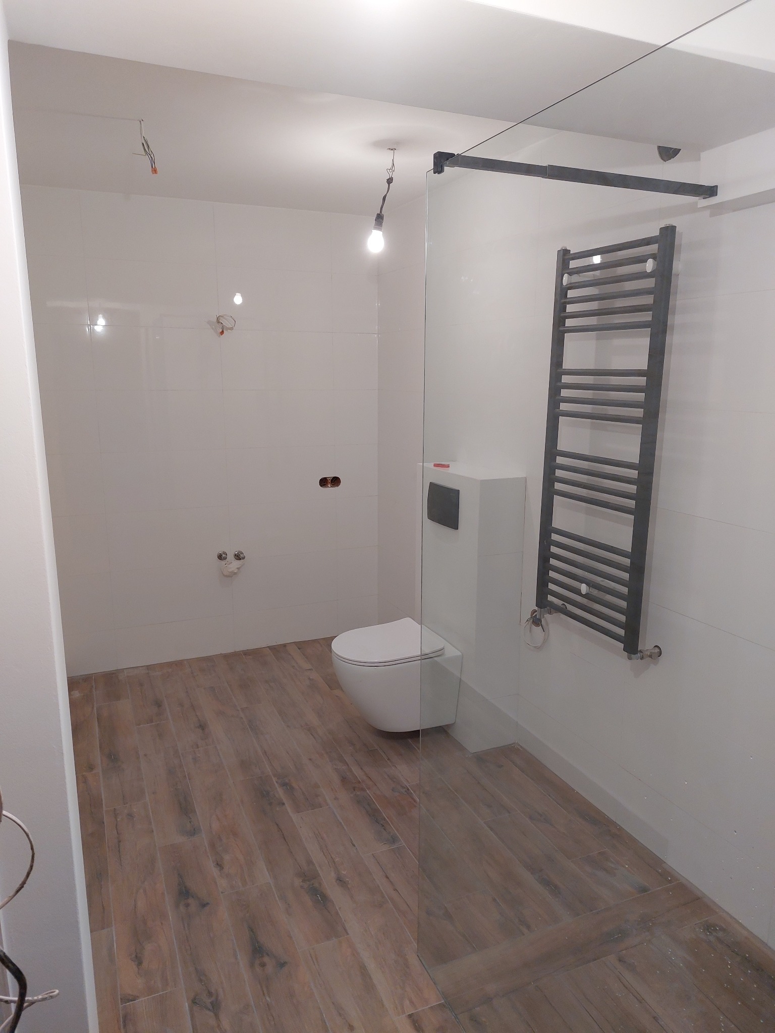 Nowoczesna łazienka z drewnopodobną podłogą, białą ceramiką i grzejnikiem drabinkowym w szarym kolorze, oddzielona szklaną ścianką, widoczne niedokończone instalacje elektryczne.