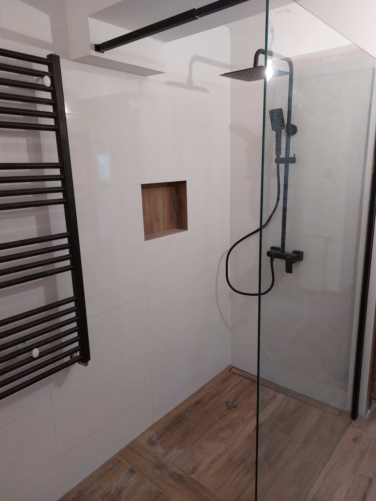 Nowoczesna kabina prysznicowa z czarną armaturą, szklaną ścianką i drewnopodobną podłogą, widoczny grzejnik drabinkowy i wnęka w ścianie.