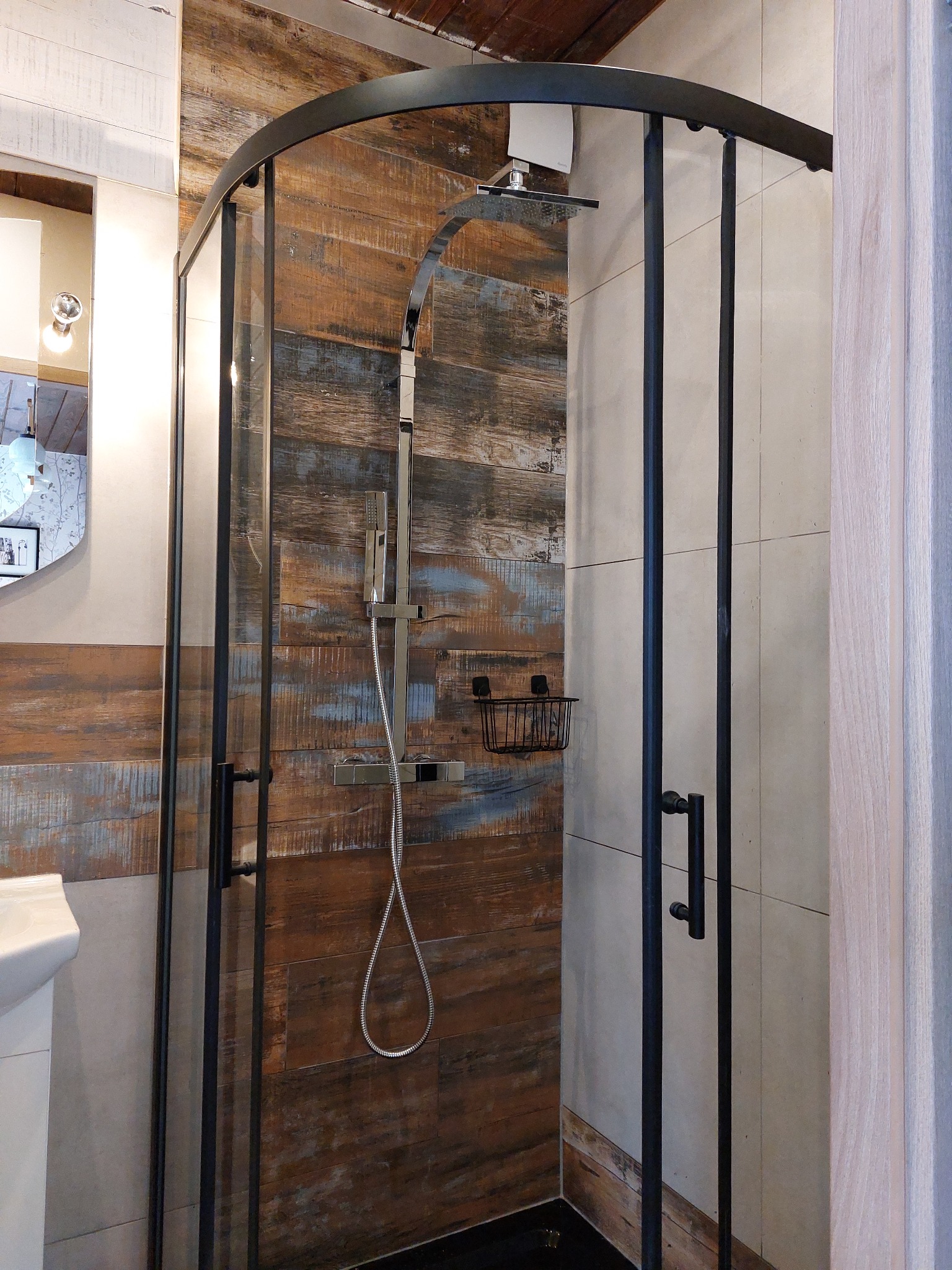 Kabina prysznicowa z czarnymi profilami na tle ściany wyłożonej płytkami imitującymi drewno i jasnymi płytkami ceramicznymi, widoczna bateria prysznicowa i półka na kosmetyki.