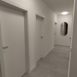 Kubatura Jakub Krzemiński - Nowoczesny korytarz w jasnych barwach z trzema białymi drzwiami, szarą podłogą z płytek i owalnym lustrem na końcu.