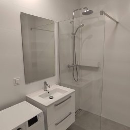 Kubatura Jakub Krzemiński - Nowoczesna, minimalistyczna łazienka z kabiną prysznicową typu walk-in, białą umywalką z szafką, lustrem i pralką, wykończona w szaro-białej kolorystyce.