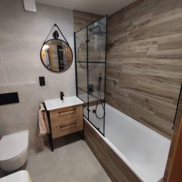 Kubatura Jakub Krzemiński - Nowoczesna łazienka z drewnopodobnymi płytkami, kabiną prysznicową typu walk-in z czarnymi profilami, okrągłym lustrem i szafką umywalkową z naturalnego drewna.