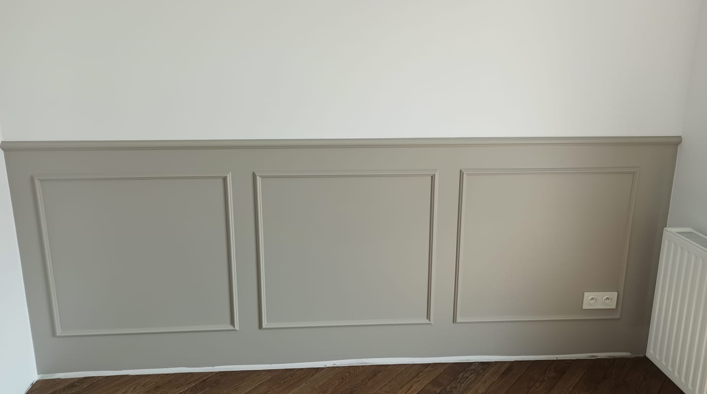 Ściana z szarymi panelami dekoracyjnymi w stylu angielskim, widoczny fragment drewnianej podłogi i biały grzejnik.