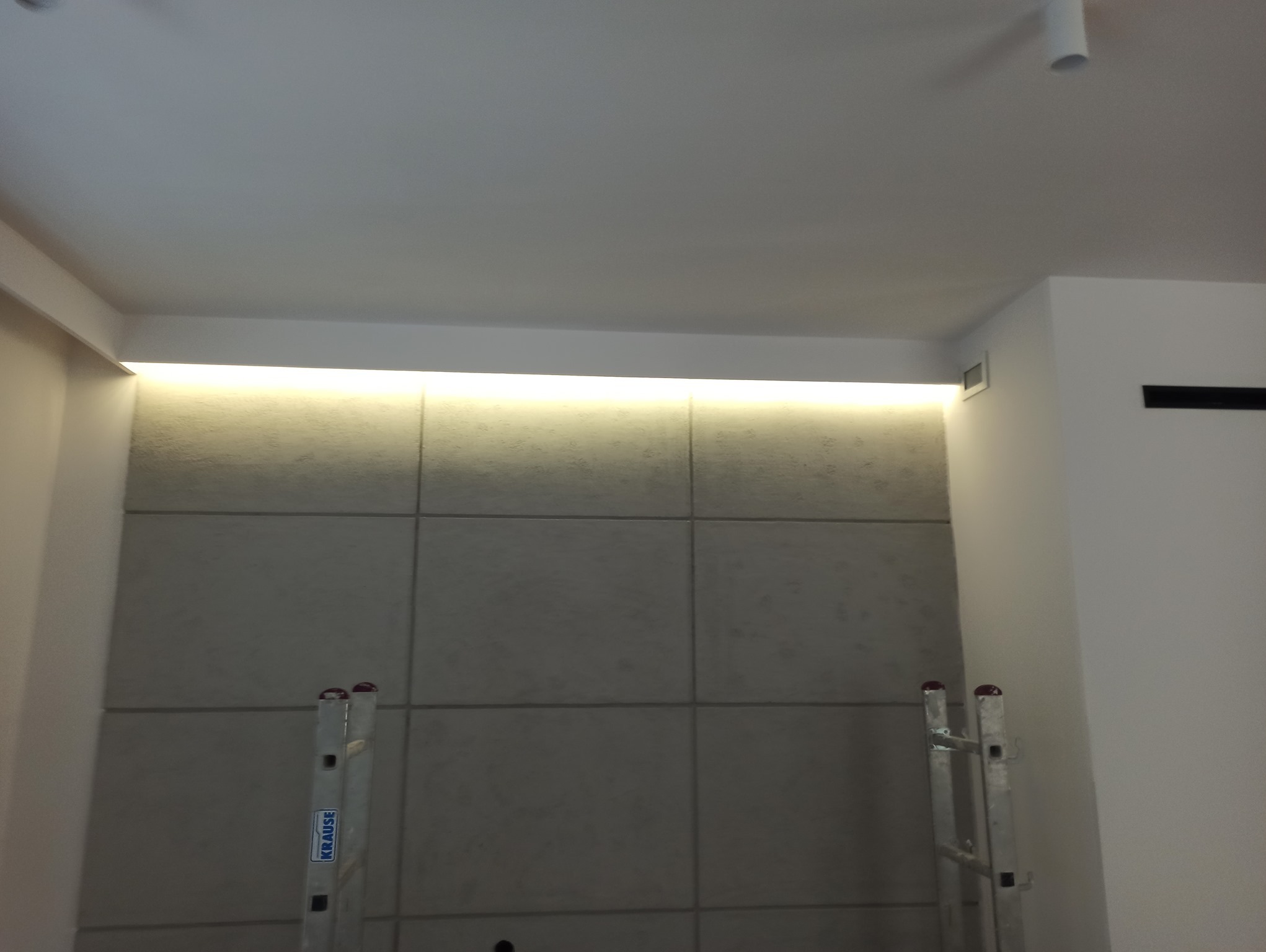 Ściana wykończona szarymi panelami z podświetleniem LED przy suficie, widoczne drabiny Krause po bokach, minimalistyczny design.