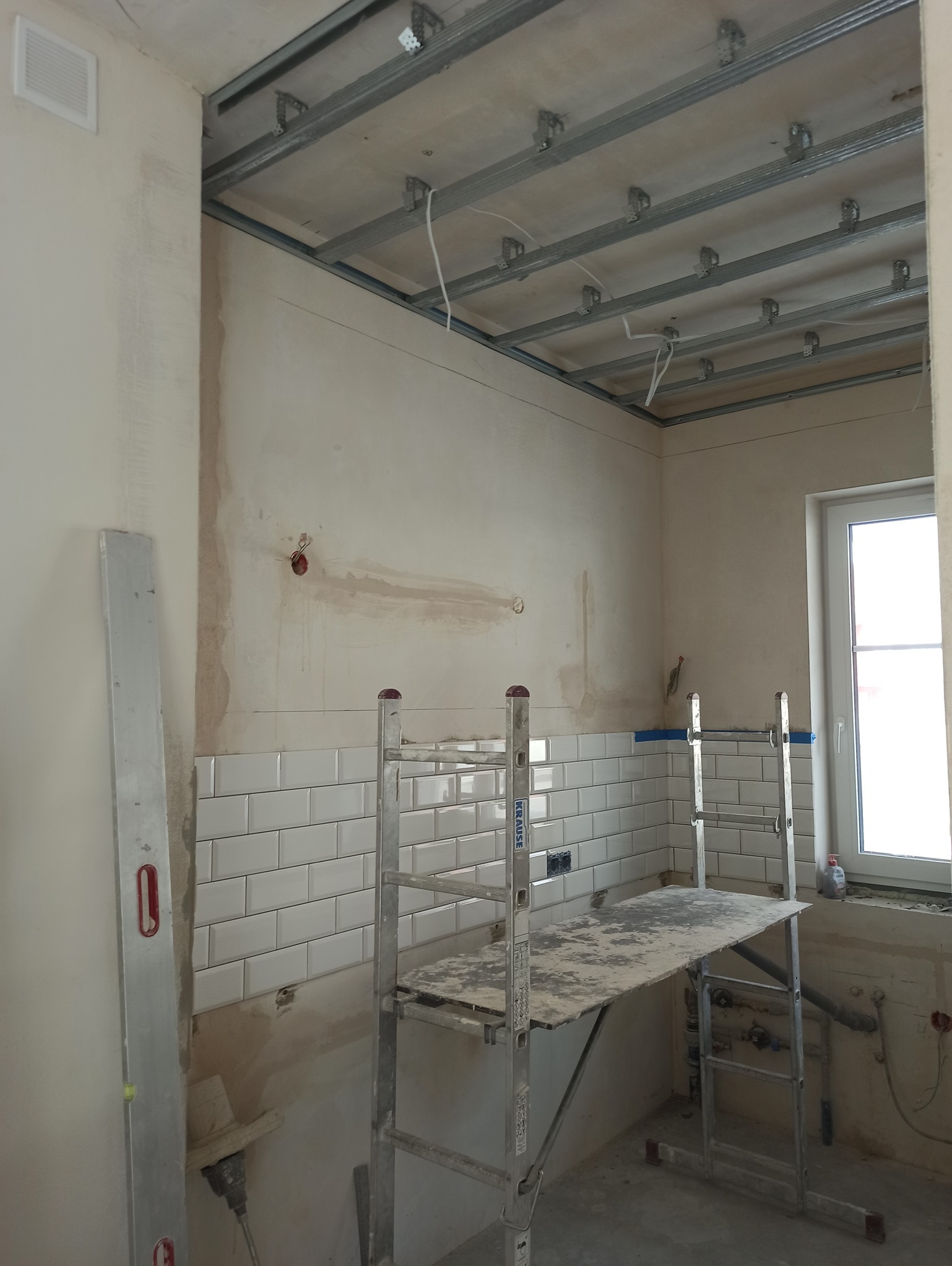 Wnętrze kuchni w trakcie prac wykończeniowych, widoczne białe płytki metra na ścianie, rusztowanie Krause, poziomica, przygotowane instalacje elektryczne i metalowa konstrukcja sufitu podwieszanego.