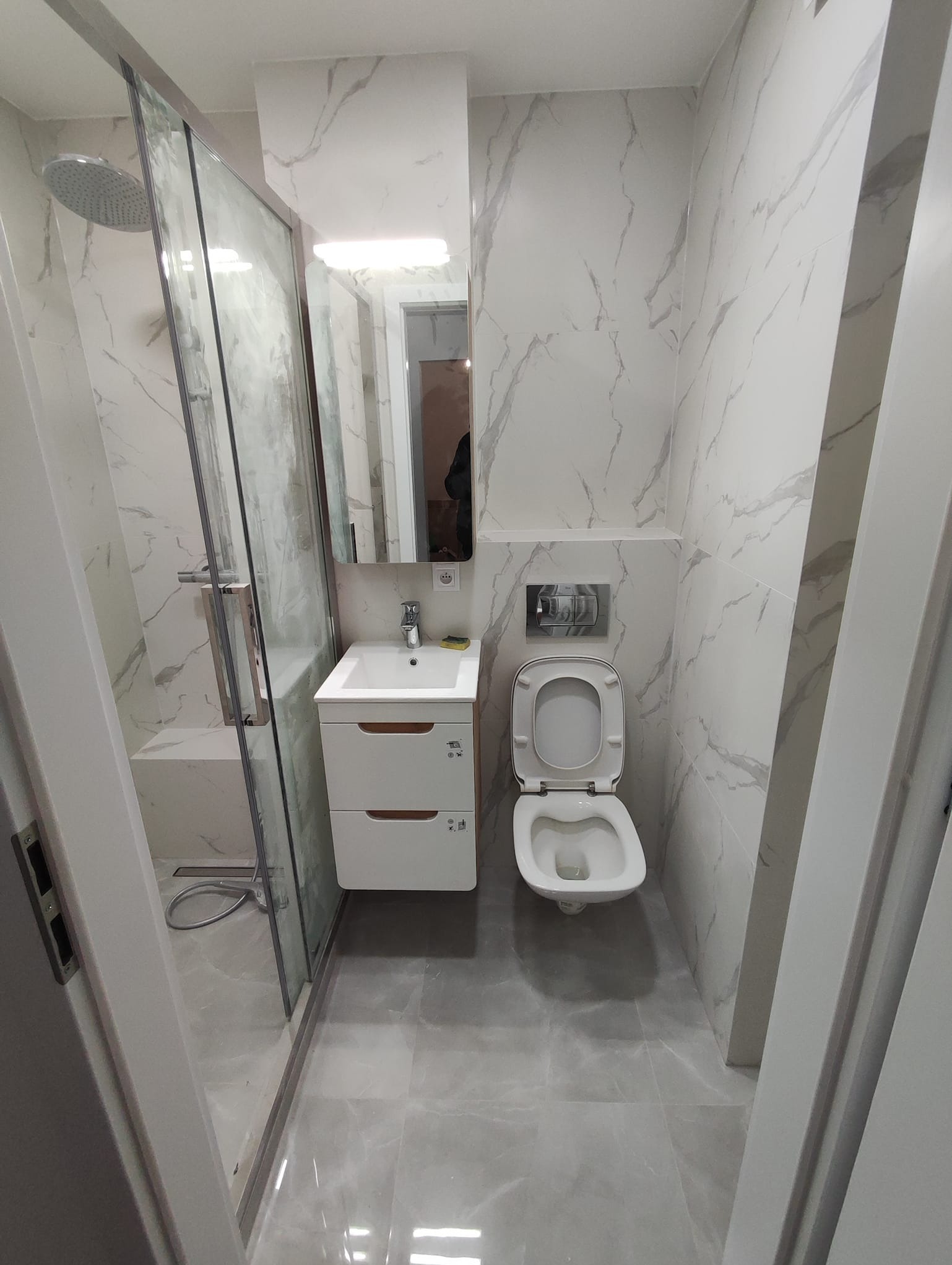 Nowoczesna łazienka z kabiną prysznicową typu walk-in, umywalką z szafką i podwieszaną toaletą, wykończona płytkami imitującymi marmur w odcieniach szarości i bieli.