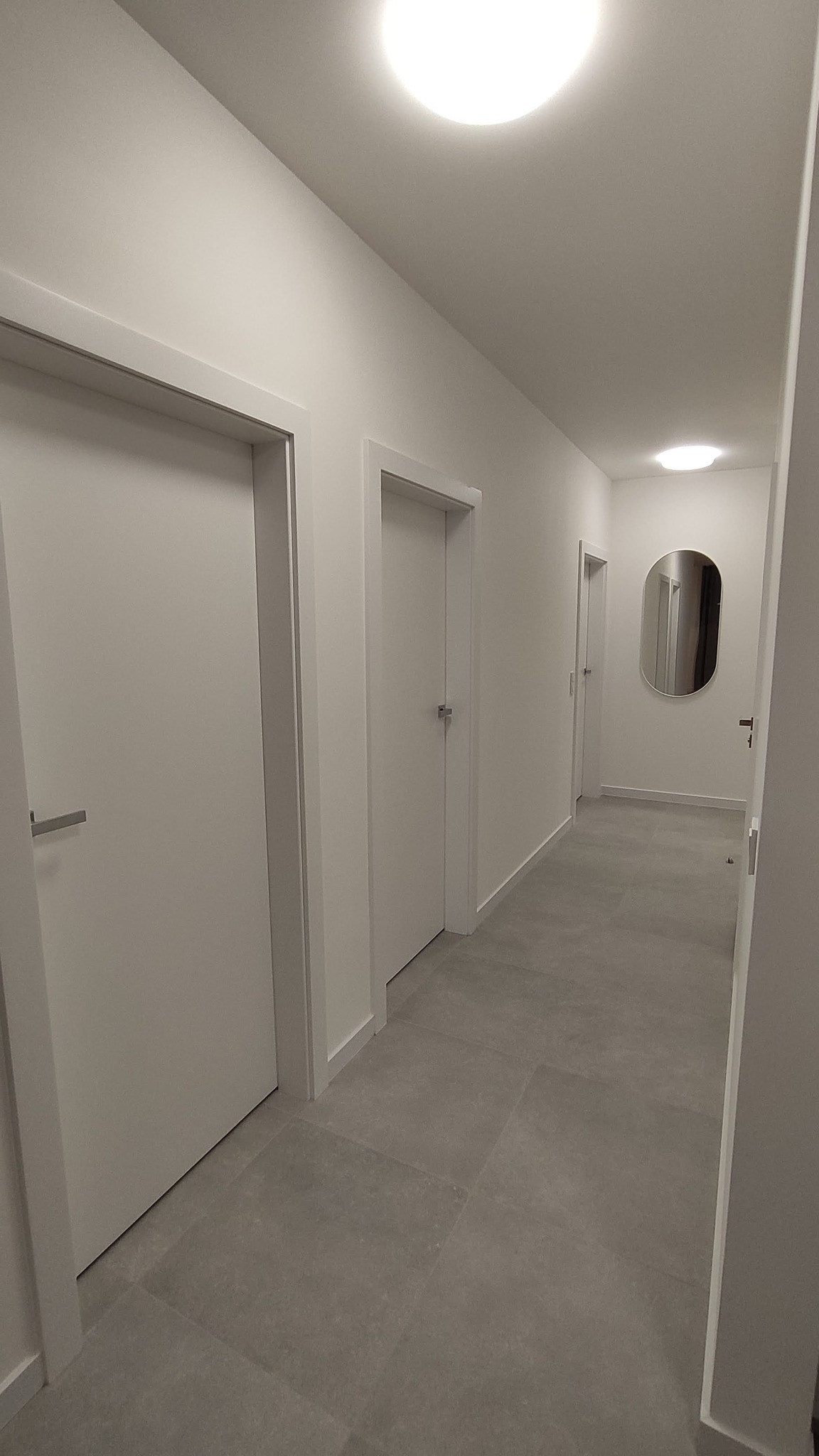 Nowoczesny korytarz w jasnych barwach z trzema białymi drzwiami, szarą podłogą z płytek i owalnym lustrem na końcu.