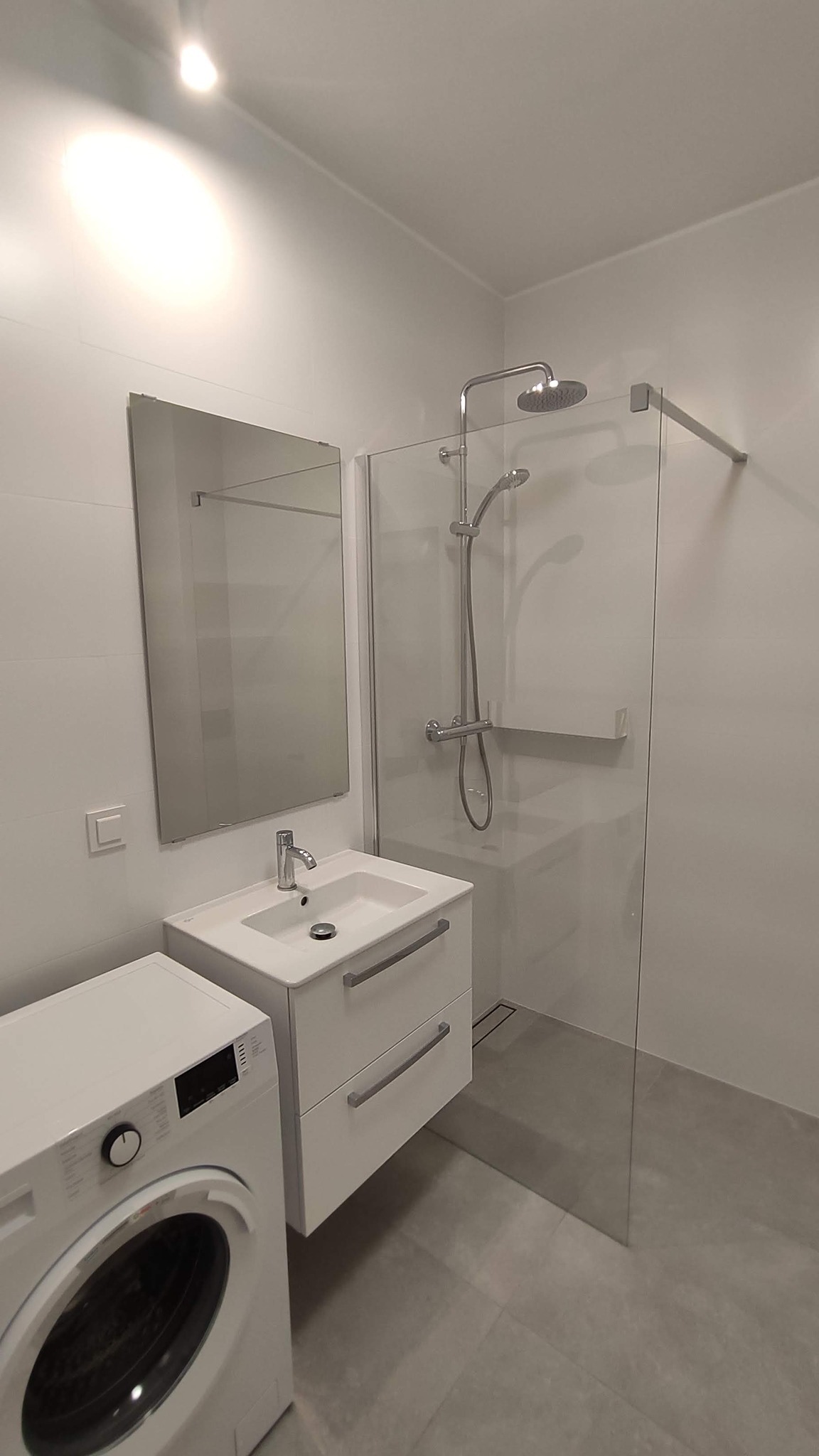 Nowoczesna, minimalistyczna łazienka z kabiną prysznicową typu walk-in, białą umywalką z szafką, lustrem i pralką, wykończona w szaro-białej kolorystyce.