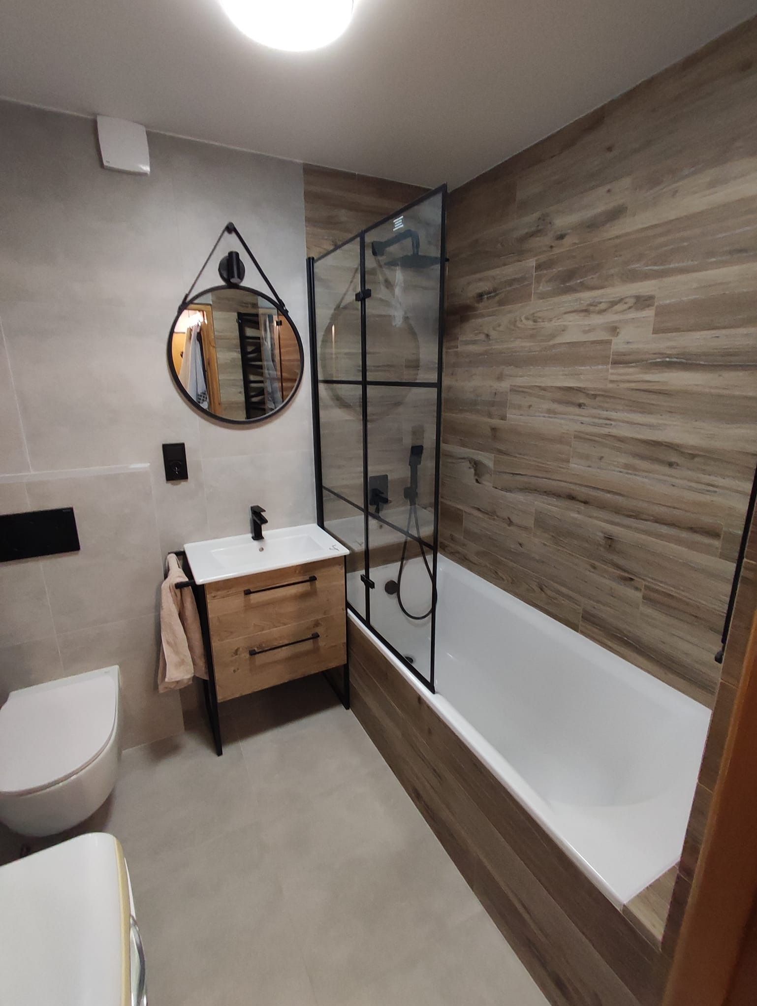 Nowoczesna łazienka z drewnopodobnymi płytkami, kabiną prysznicową typu walk-in z czarnymi profilami, okrągłym lustrem i szafką umywalkową z naturalnego drewna.