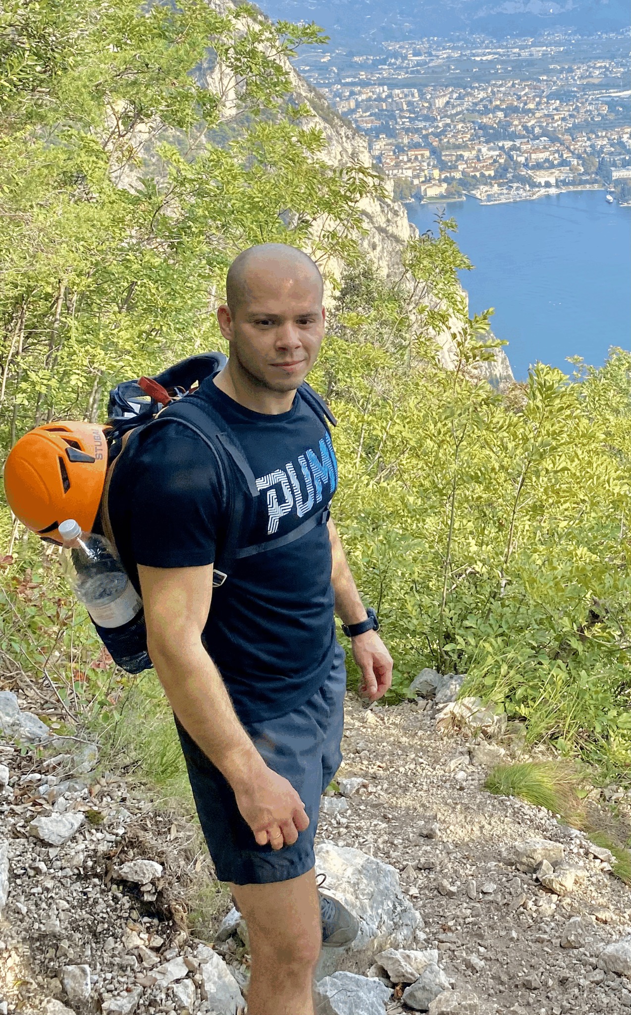 Mężczyzna z plecakiem i pomarańczowym kaskiem w górach, widok na miasto i jezioro w tle.