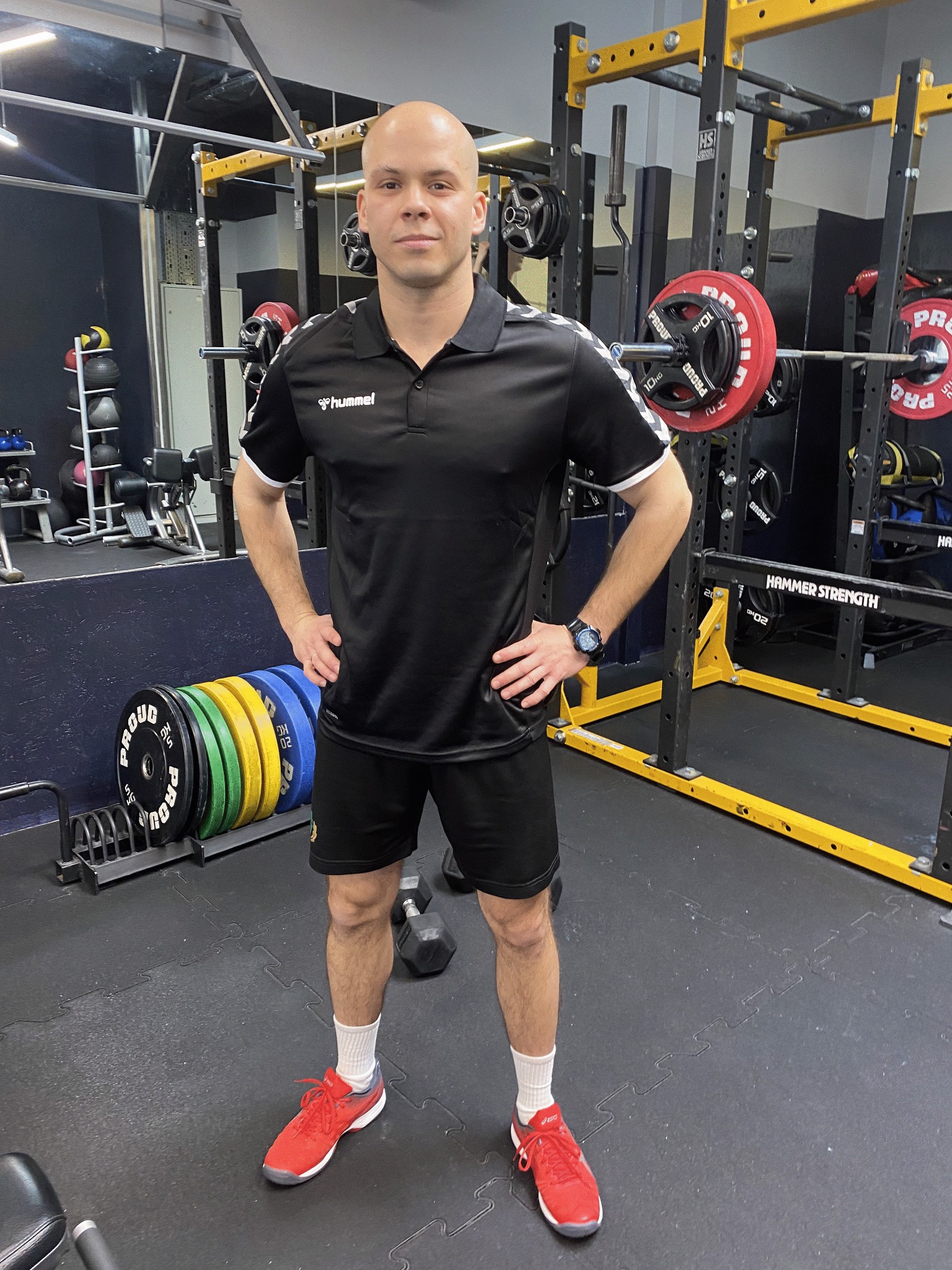 Mężczyzna w sportowym stroju Hummel stoi w siłowni na tle sprzętu do ćwiczeń siłowych, w tym sztangi, obciążenia i stojaki Hammer Strength.