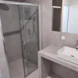 Nowoczesna łazienka z szarą glazurą, przeszkloną kabiną prysznicową z deszczownicą i białą prostokątną umywalką nablatową na betonowym blacie.