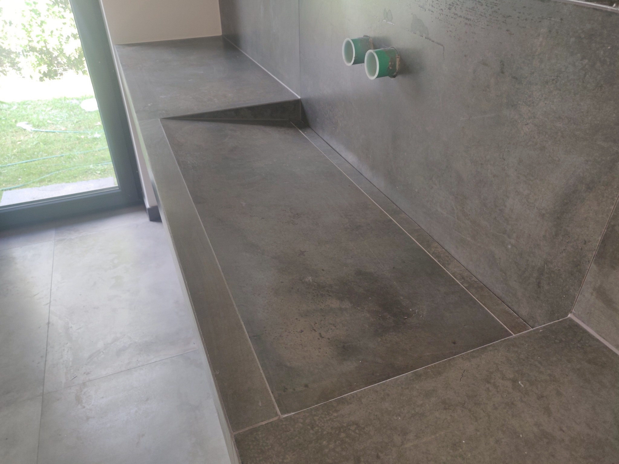 Nowoczesna, ciemna, kamienna umywalka w łazience, z widocznymi rurami doprowadzającymi wodę w ścianie i oknem z widokiem na ogród.