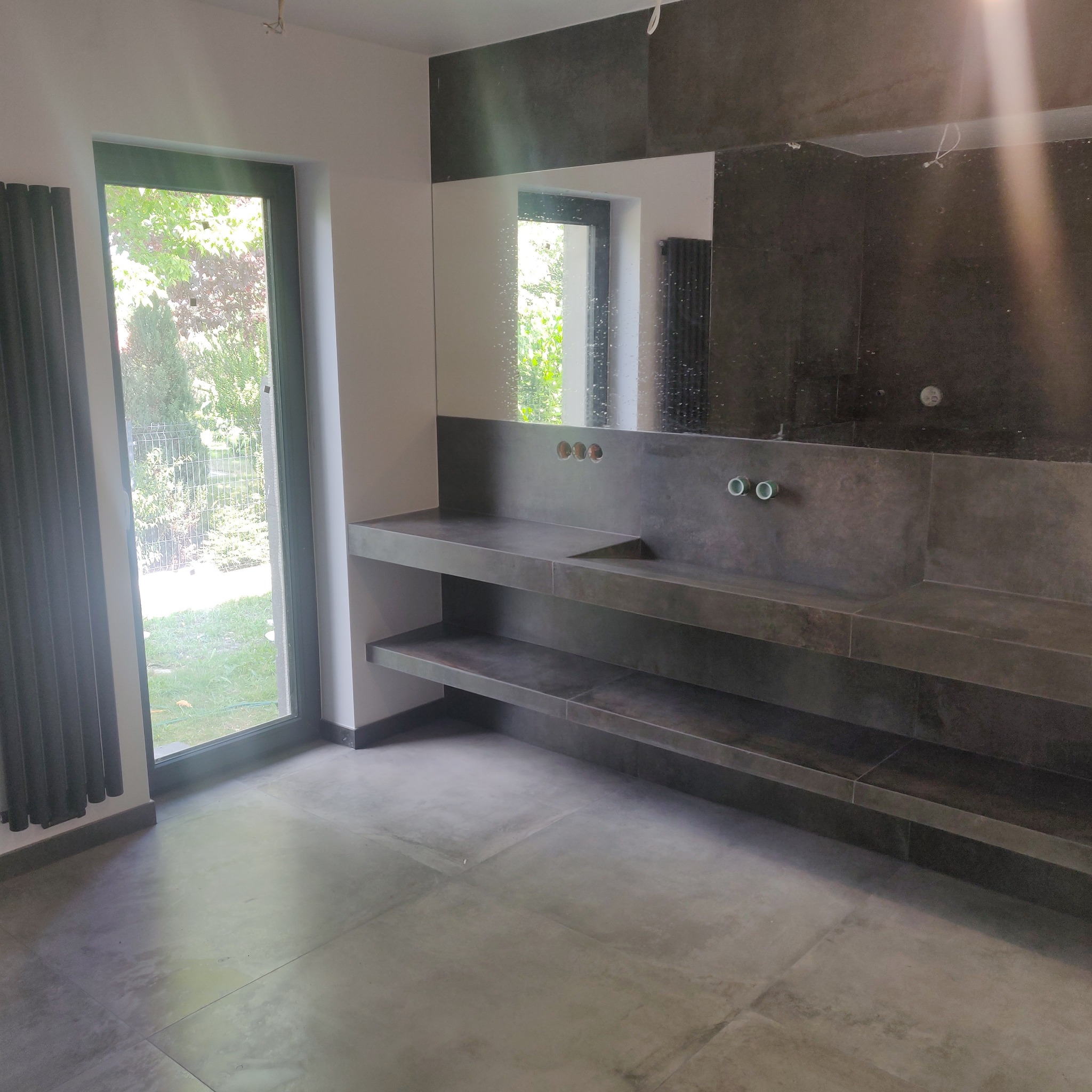 Nowoczesna łazienka w Bolesławcu z szarą, betonową zabudową umywalki z półkami, dużym lustrem i szarymi płytkami podłogowymi, widok na zieleń przez okno.