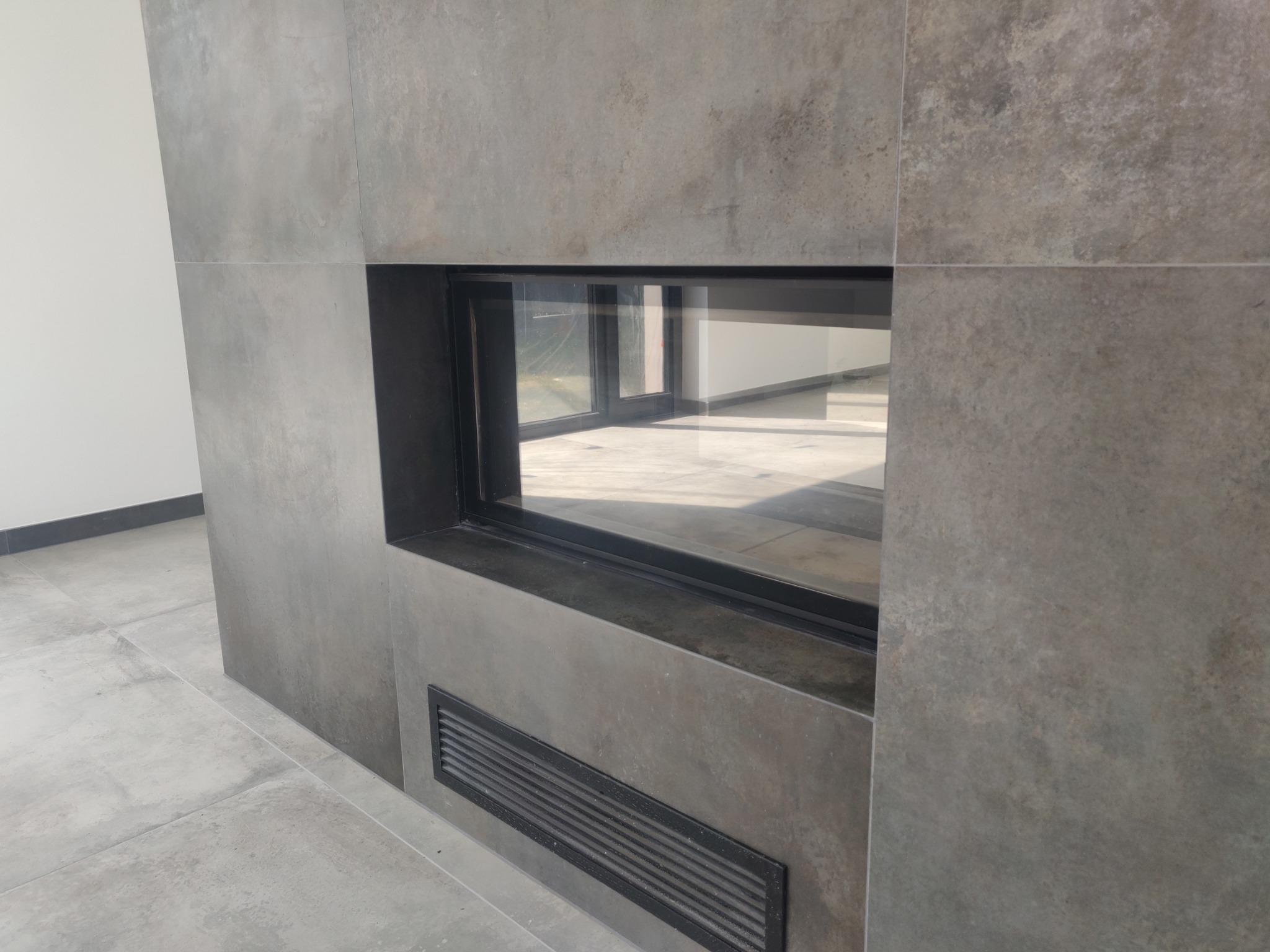 Wykończony kominek z szarą okładziną imitującą beton, z czarną ramą i kratką wentylacyjną u dołu, widok z boku na tle jasnej ściany i podłogi z szarych płytek.