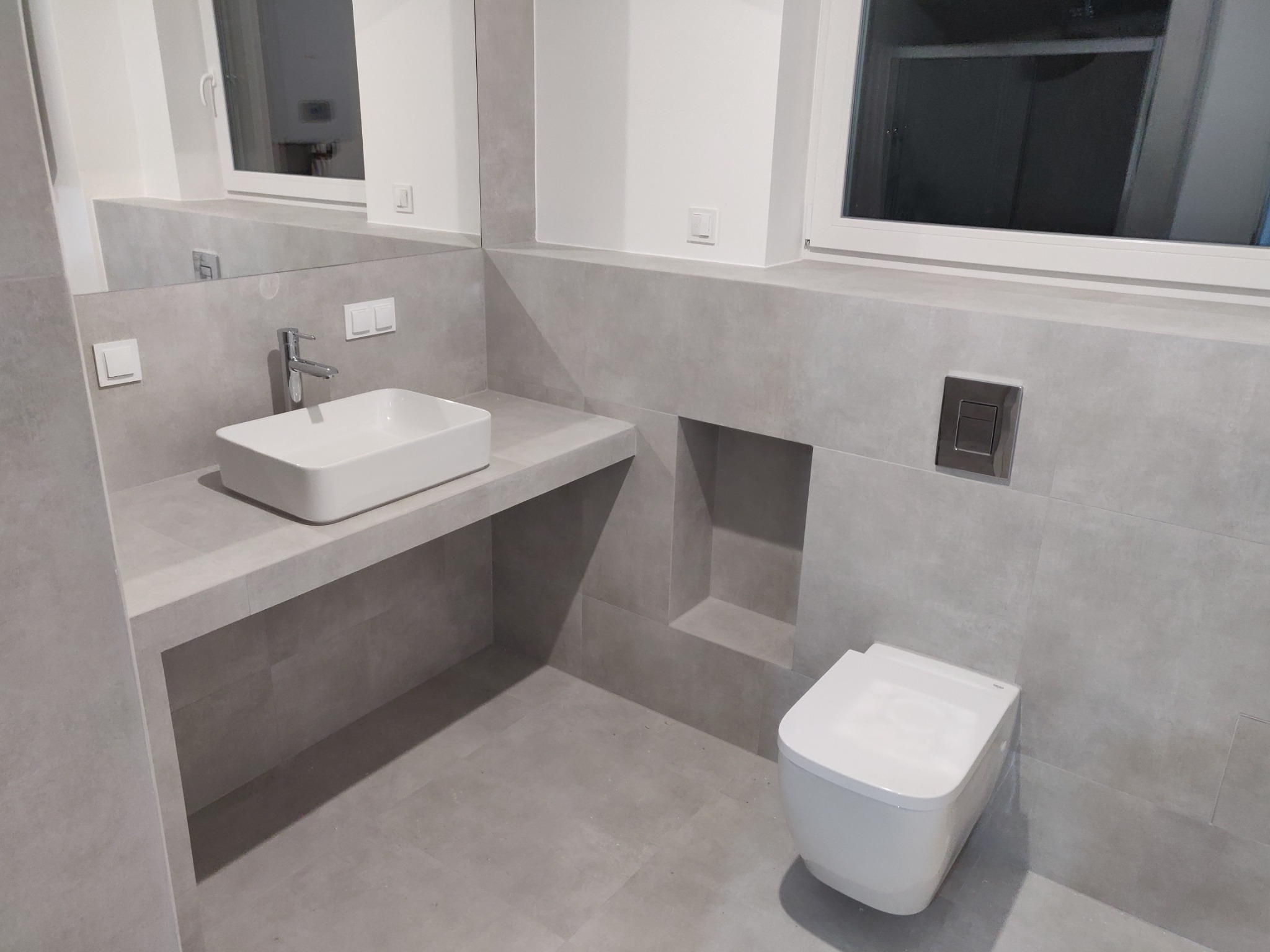 Nowoczesna łazienka w odcieniach szarości, z podwieszaną toaletą i umywalką nablatową na kamiennym blacie, minimalistyczna armatura, widoczne płytki imitujące kamień na ścianach i podłodze...