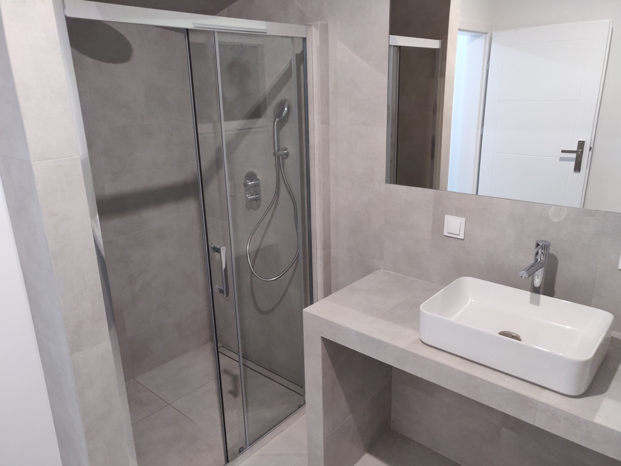 Nowoczesna łazienka z szarą glazurą, przeszkloną kabiną prysznicową z deszczownicą i białą prostokątną umywalką nablatową na betonowym blacie.