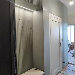 Arkomeble - Wnęka z siedziskiem i wieszakami, oświetlona listwą LED, z pionowymi panelami na ścianie, obok białe drzwi wewnętrzne, widok na remontowane pomieszczenie.