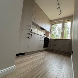 Arkomeble - Jasnoszara zabudowa kuchenna z drewnianym blatem, czarnymi uchwytami i oknem z widokiem na drzewo, oświetlona reflektorami na suficie, z podłogą imitującą drewno.