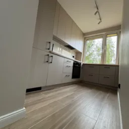 Jasnoszara zabudowa kuchenna z drewnianym blatem, czarnymi uchwytami i oknem z widokiem na drzewo, oświetlona reflektorami na suficie, z podłogą imitującą drewno.