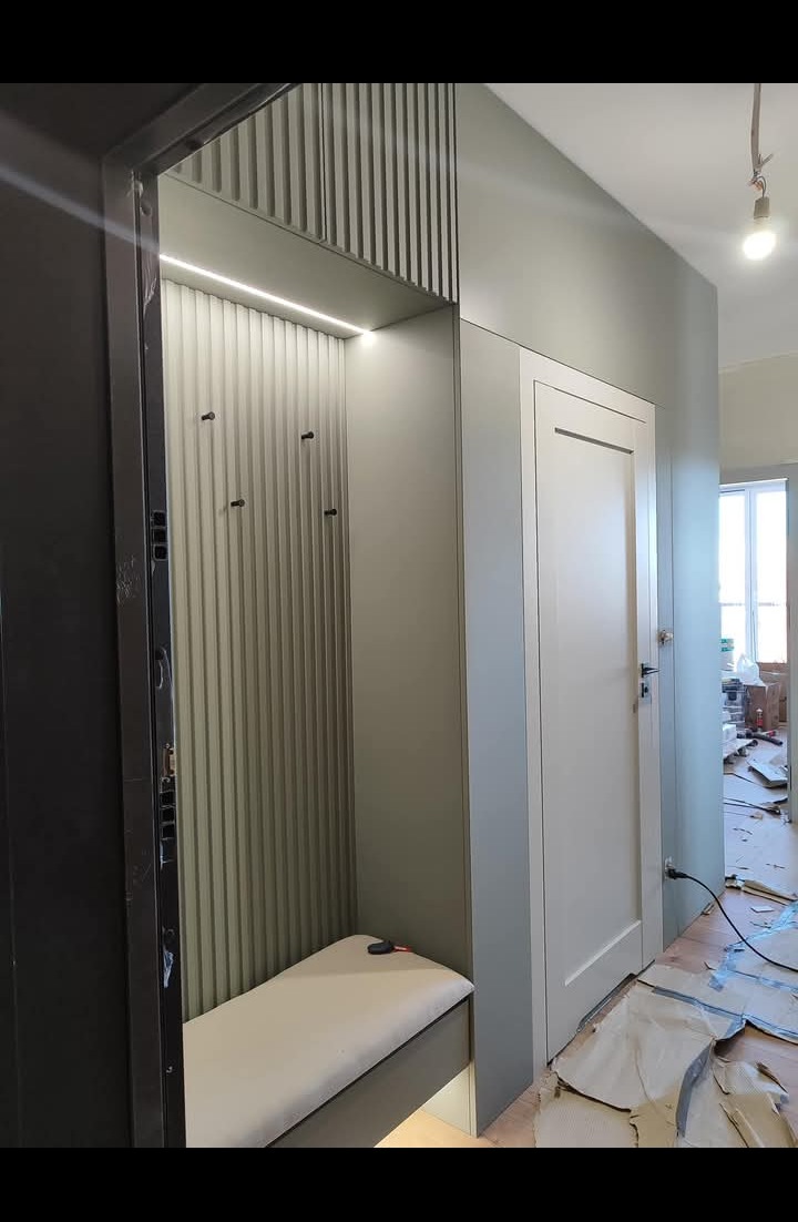 Wnęka z siedziskiem i wieszakami, oświetlona listwą LED, z pionowymi panelami na ścianie, obok białe drzwi wewnętrzne, widok na remontowane pomieszczenie.