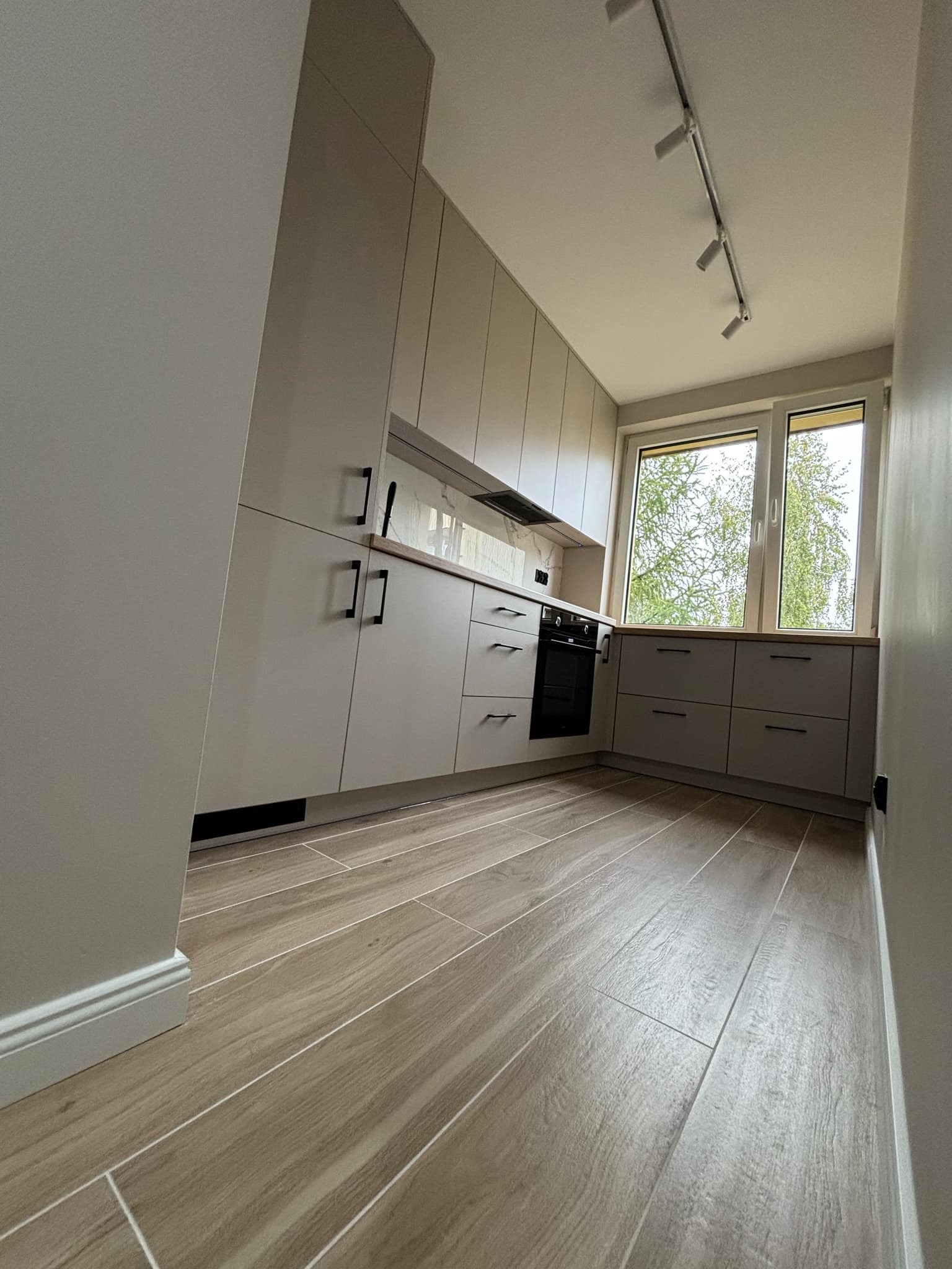 Jasnoszara zabudowa kuchenna z drewnianym blatem, czarnymi uchwytami i oknem z widokiem na drzewo, oświetlona reflektorami na suficie, z podłogą imitującą drewno.