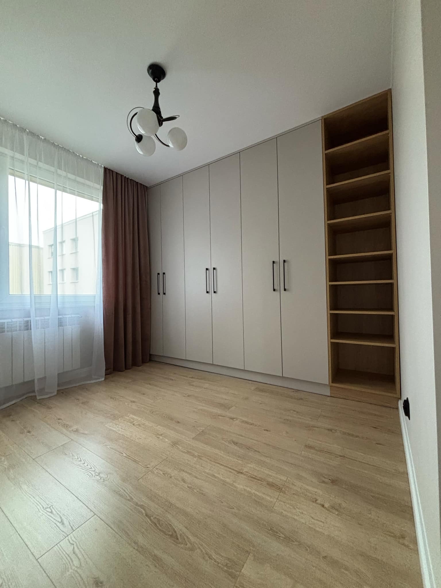 Jasnoszara szafa wnękowa z czarnymi uchwytami i otwartym regałem z jasnego drewna, oświetlona przez okno z białymi i brązowymi zasłonami, uzupełniona drewnopodobną podłogą.