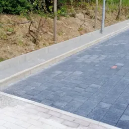 Ułożona nawierzchnia z kostki brukowej w odcieniach szarości i czerwieni, obrzeża z jasnego betonu, widoczny fragment ławki i zieleń w tle.