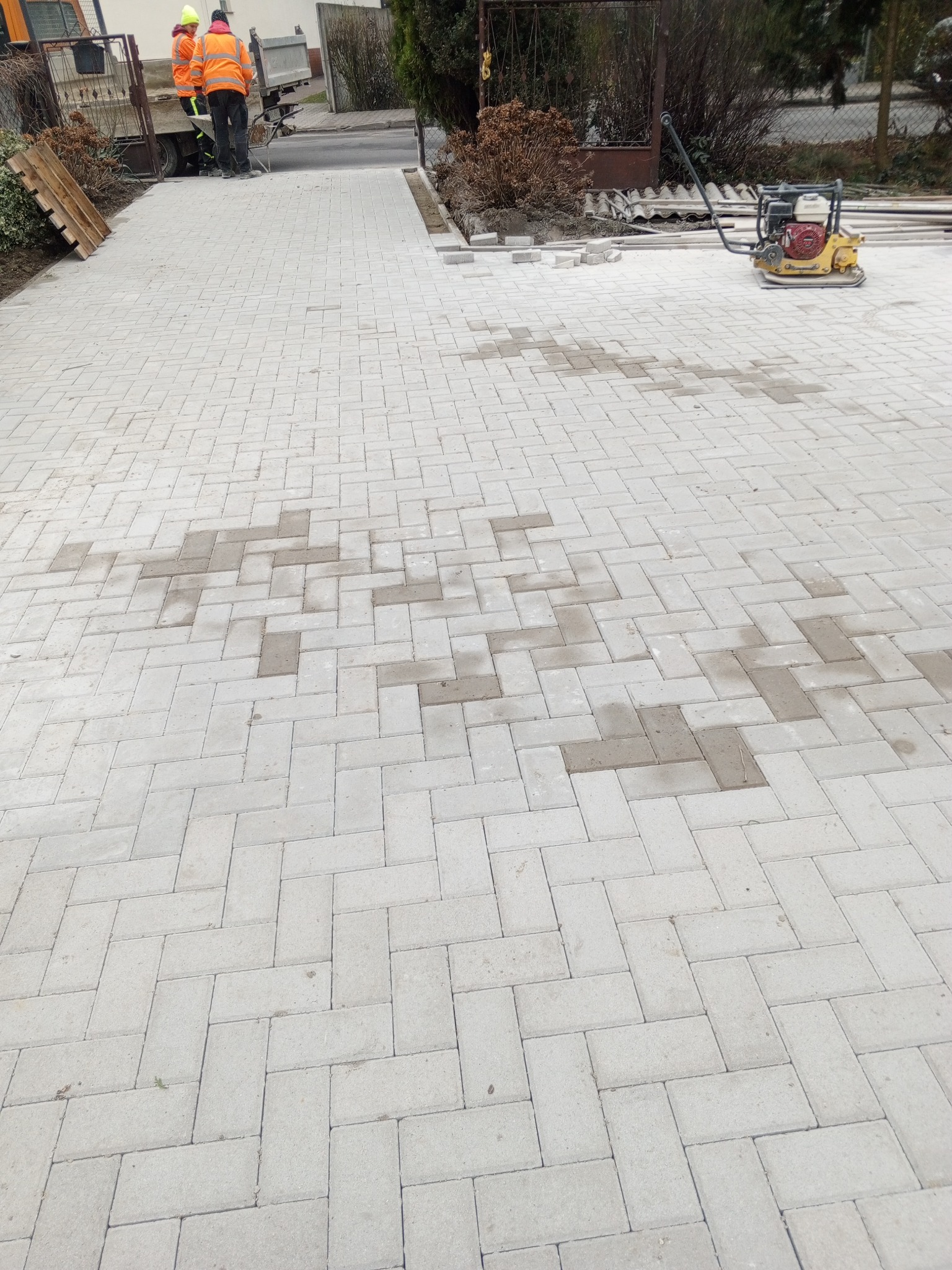 Ułożona, jasnoszara kostka brukowa w jodełkę na podjeździe, z widocznymi plamami i zagęszczarką spalinową w tle oraz pracownikami przy ciężarówce.