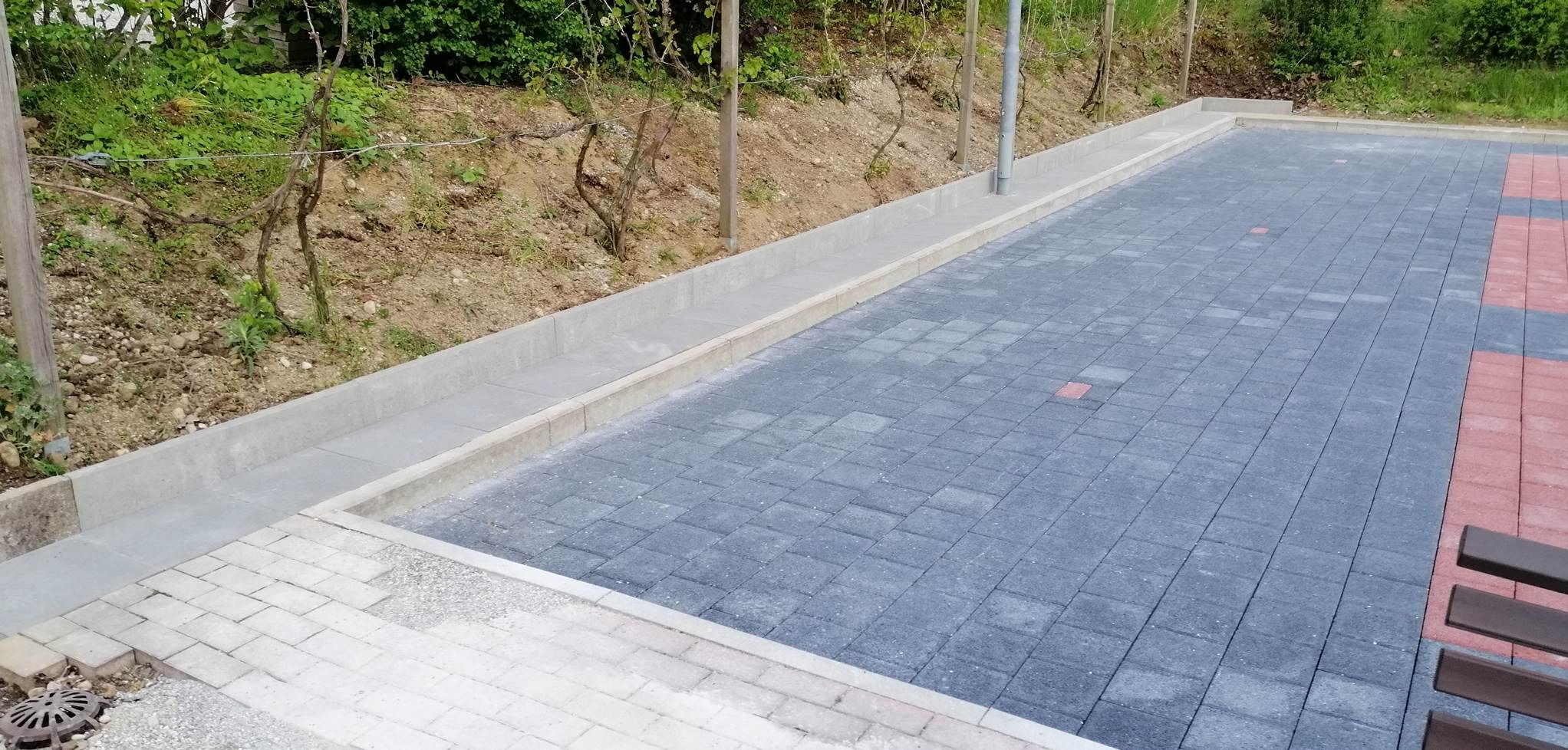 Ułożona nawierzchnia z kostki brukowej w odcieniach szarości i czerwieni, obrzeża z jasnego betonu, widoczny fragment ławki i zieleń w tle.