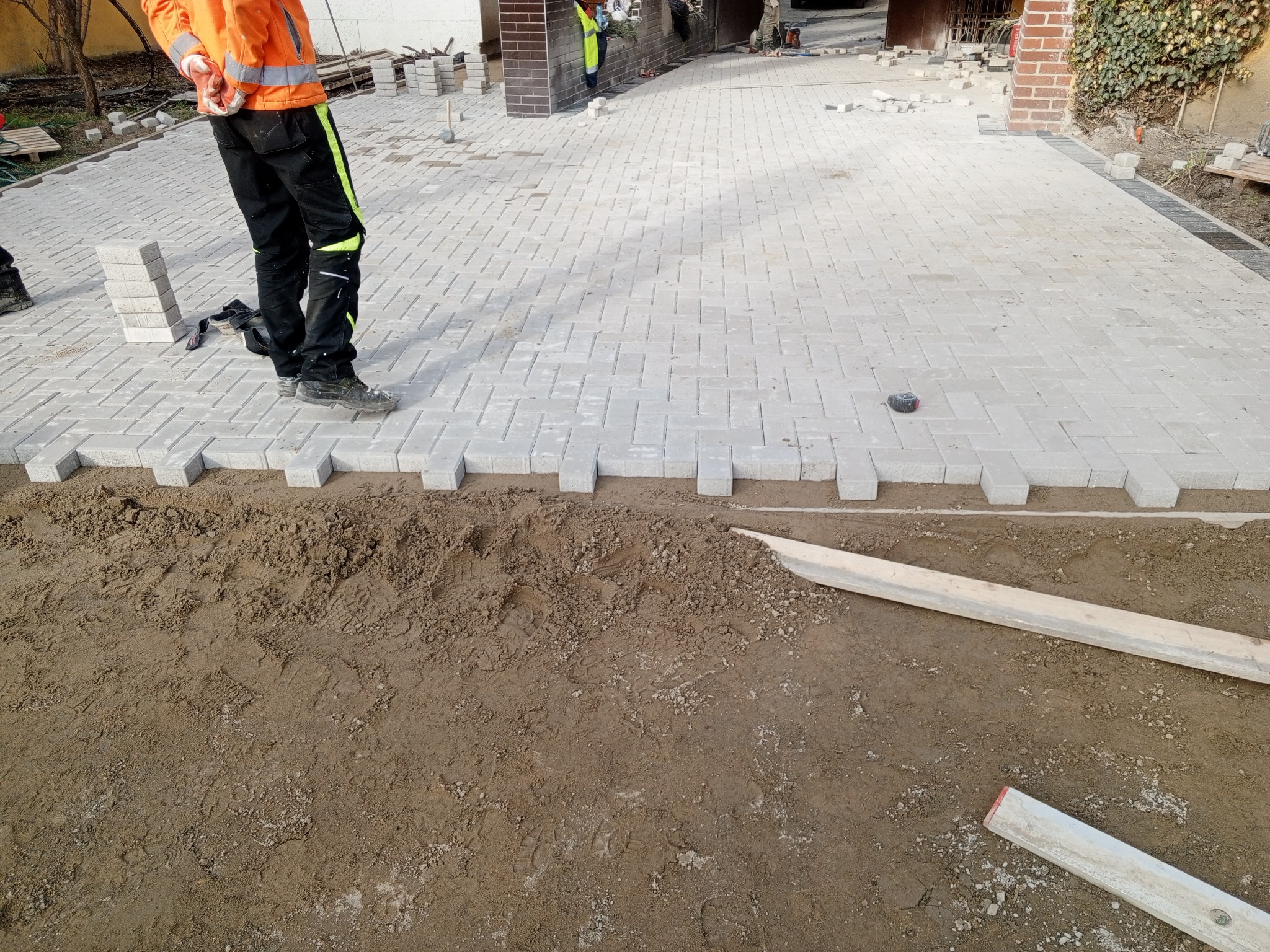 Ułożona częściowo szara kostka brukowa w jodełkę na posesji, widoczny pracownik w odblaskowej kamizelce i przygotowane podłoże z ziemi.