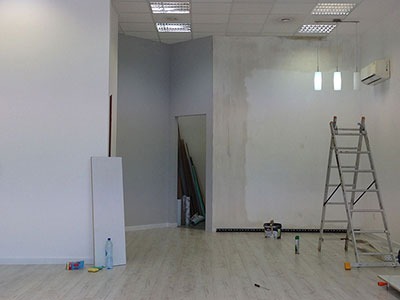 Pokój w trakcie prac wykończeniowych, z częściowo wygładzoną ścianą, drabiną, narzędziami i listwami przypodłogowymi w tle.