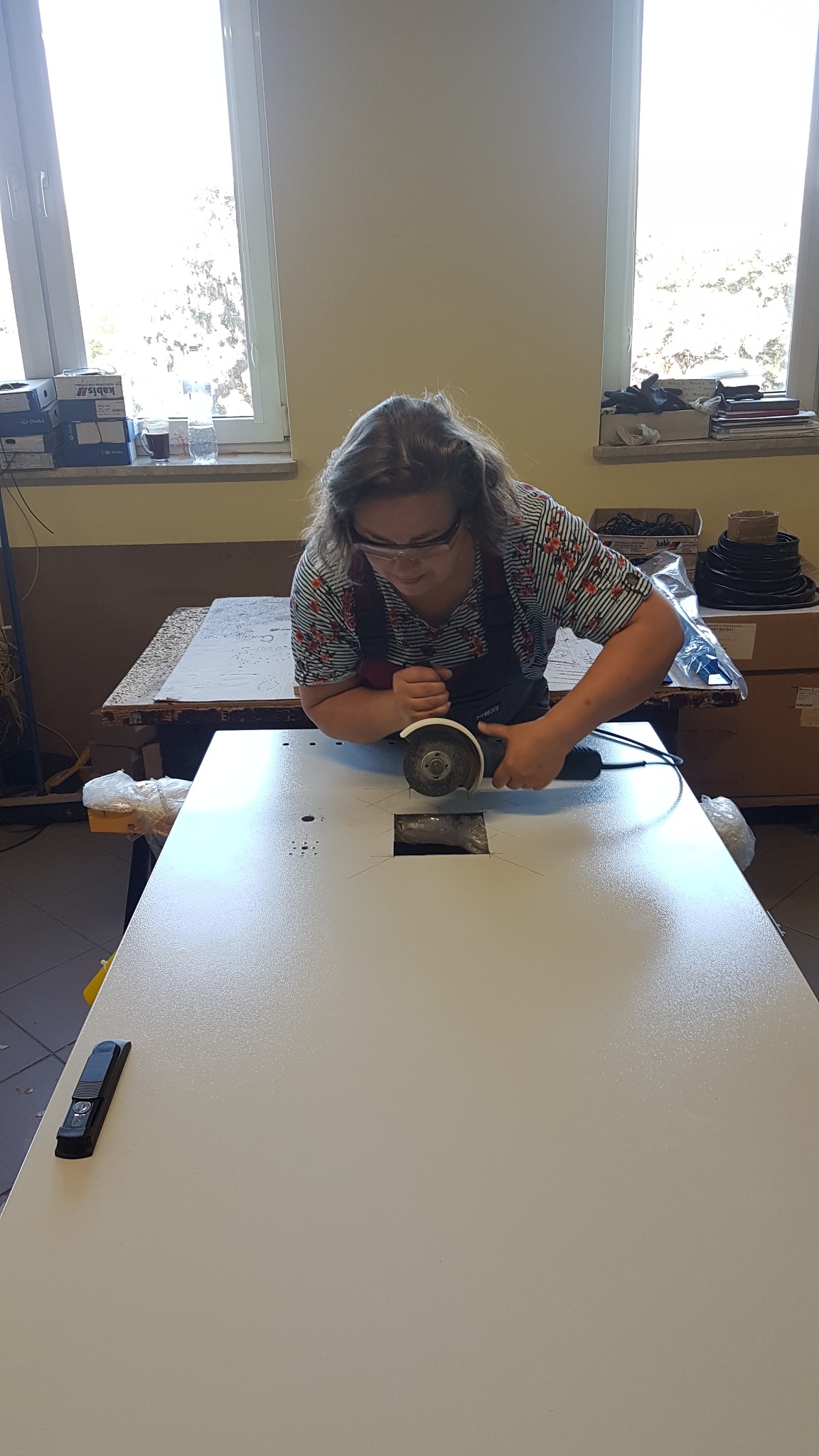 Pracownik w okularach ochronnych wycina otwór szlifierką kątową w metalowej obudowie elektrycznej, widoczne oznaczenia linii cięcia.