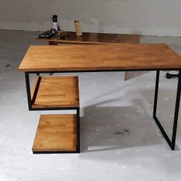 FransWeld Franciszek Nowak - Stół z drewnianym blatem i metalową ramą z półkami w surowym wnętrzu, widoczna piła ręczna i lampa budowlana.