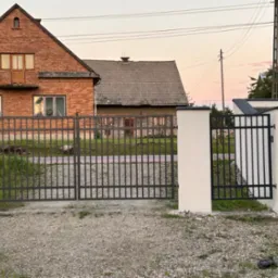 Nowoczesna, prosta brama wjazdowa z metalowymi prętami, osadzona pomiędzy białymi, murowanymi słupkami na tle ceglanego domu z drewnianymi elementami i żwirowego podjazdu.