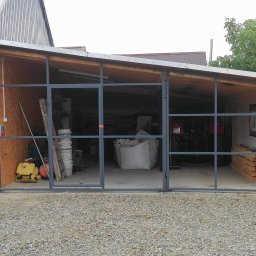 FransWeld Franciszek Nowak - Metalowa konstrukcja szkieletowa wiaty garażowej z widocznym wyposażeniem wewnątrz, takim jak traktor i materiały budowlane, z posadzką żwirową na zewnątrz.