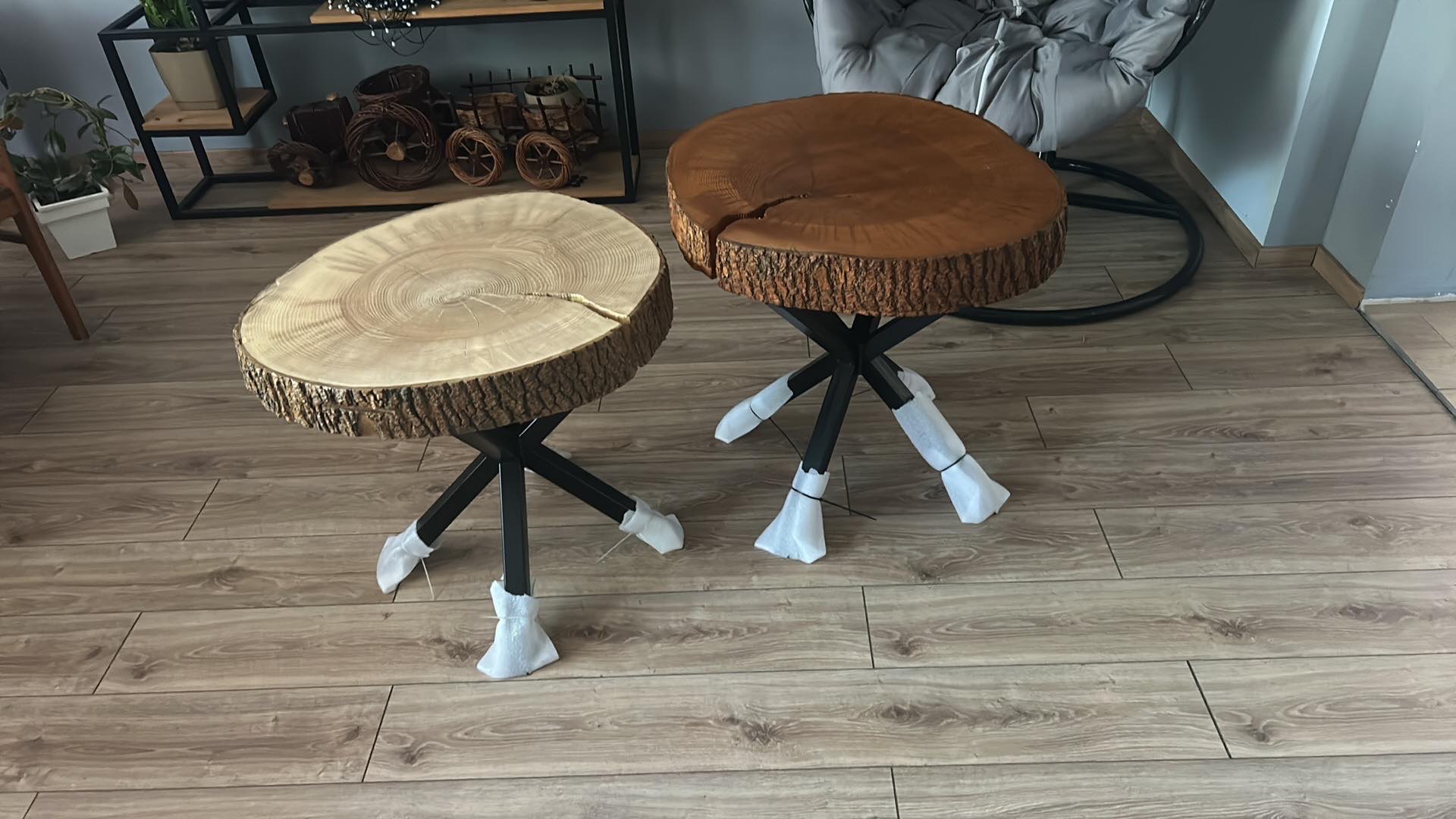 Dwa stoliki kawowe z blatami z plastrów drewna o naturalnej korze, na metalowych nogach z zabezpieczeniami transportowymi, ustawione na podłodze z paneli.