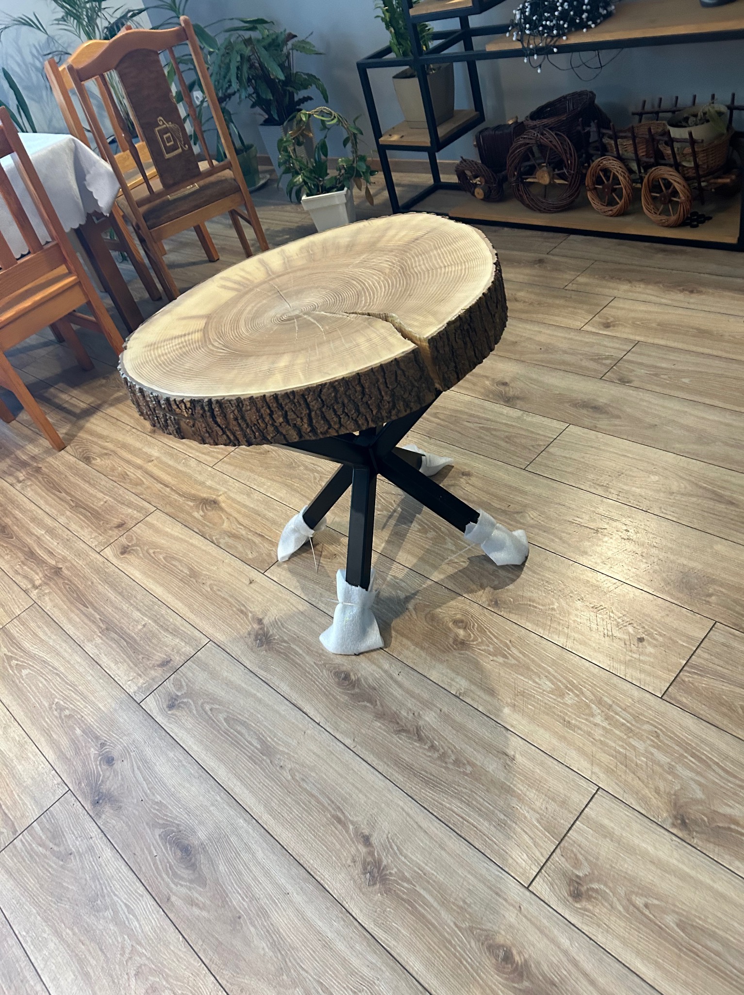 Stolik kawowy z blatem z plastra drewna o naturalnej korze, osadzony na czarnych, metalowych nogach z zabezpieczeniami na końcach, na tle drewnianej podłogi i wyposażenia wnętrza.