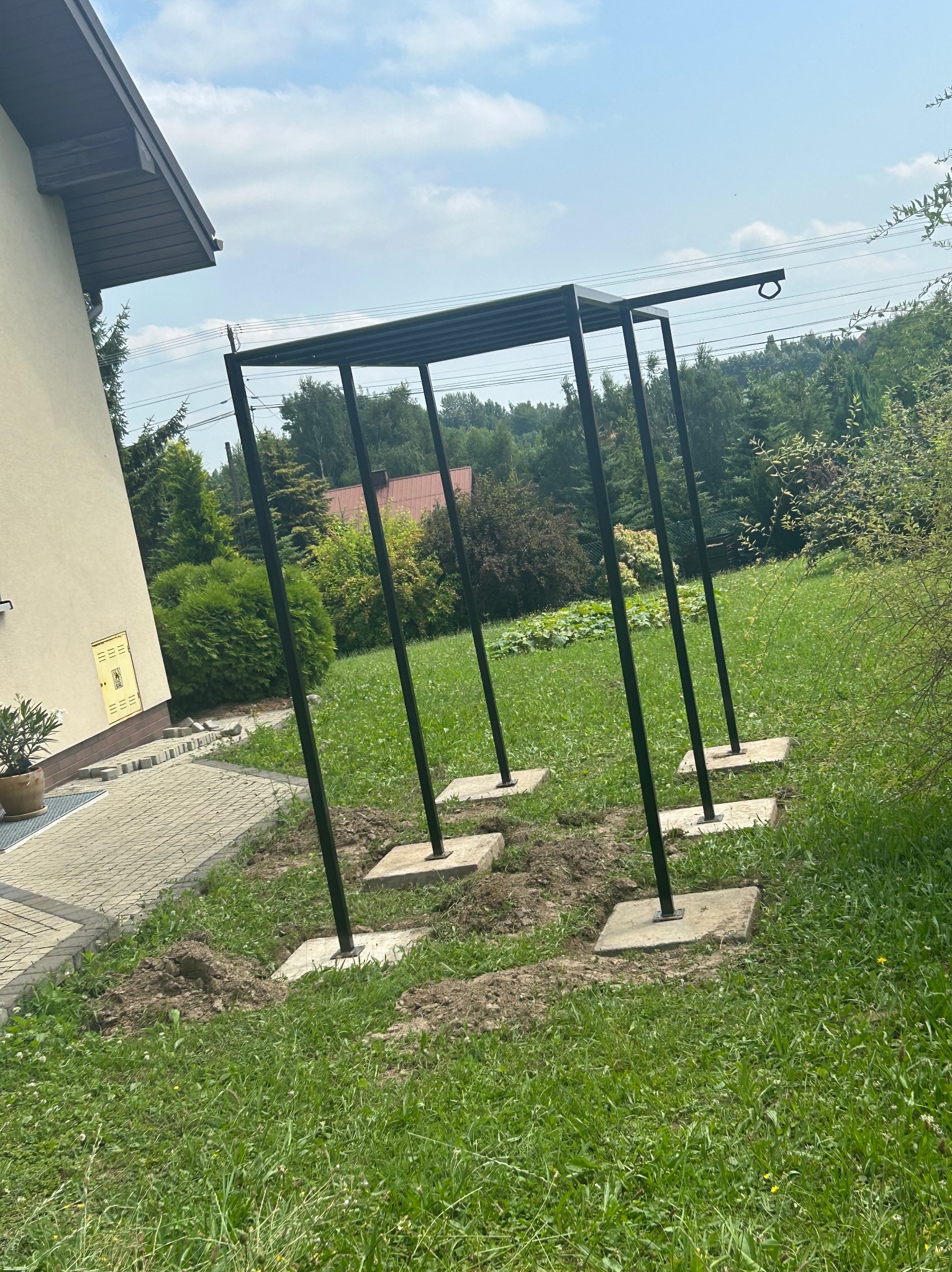 Czarna, metalowa konstrukcja z czterema słupami na betonowych podstawach, prawdopodobnie w trakcie montażu, na trawniku przed domem jednorodzinnym w słoneczny dzień.