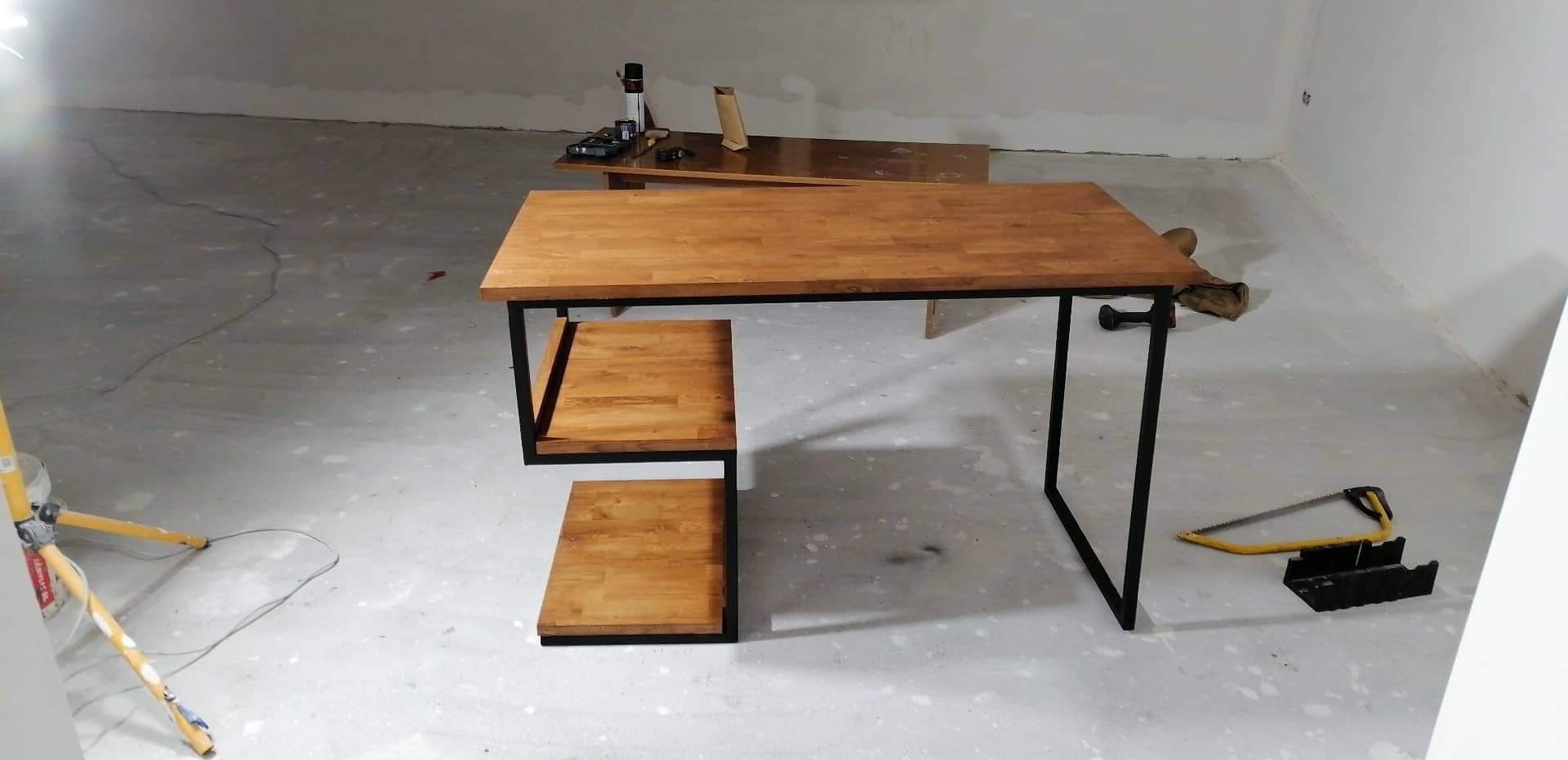 Stół z drewnianym blatem i metalową ramą z półkami w surowym wnętrzu, widoczna piła ręczna i lampa budowlana.