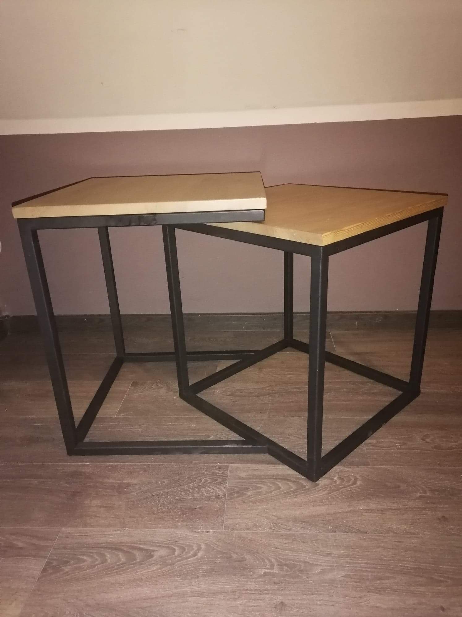 Dwa stoliki kawowe z drewnianymi blatami i czarnymi metalowymi podstawami w kształcie prostopadłościanów, ustawione jeden pod drugim na drewnianej podłodze.