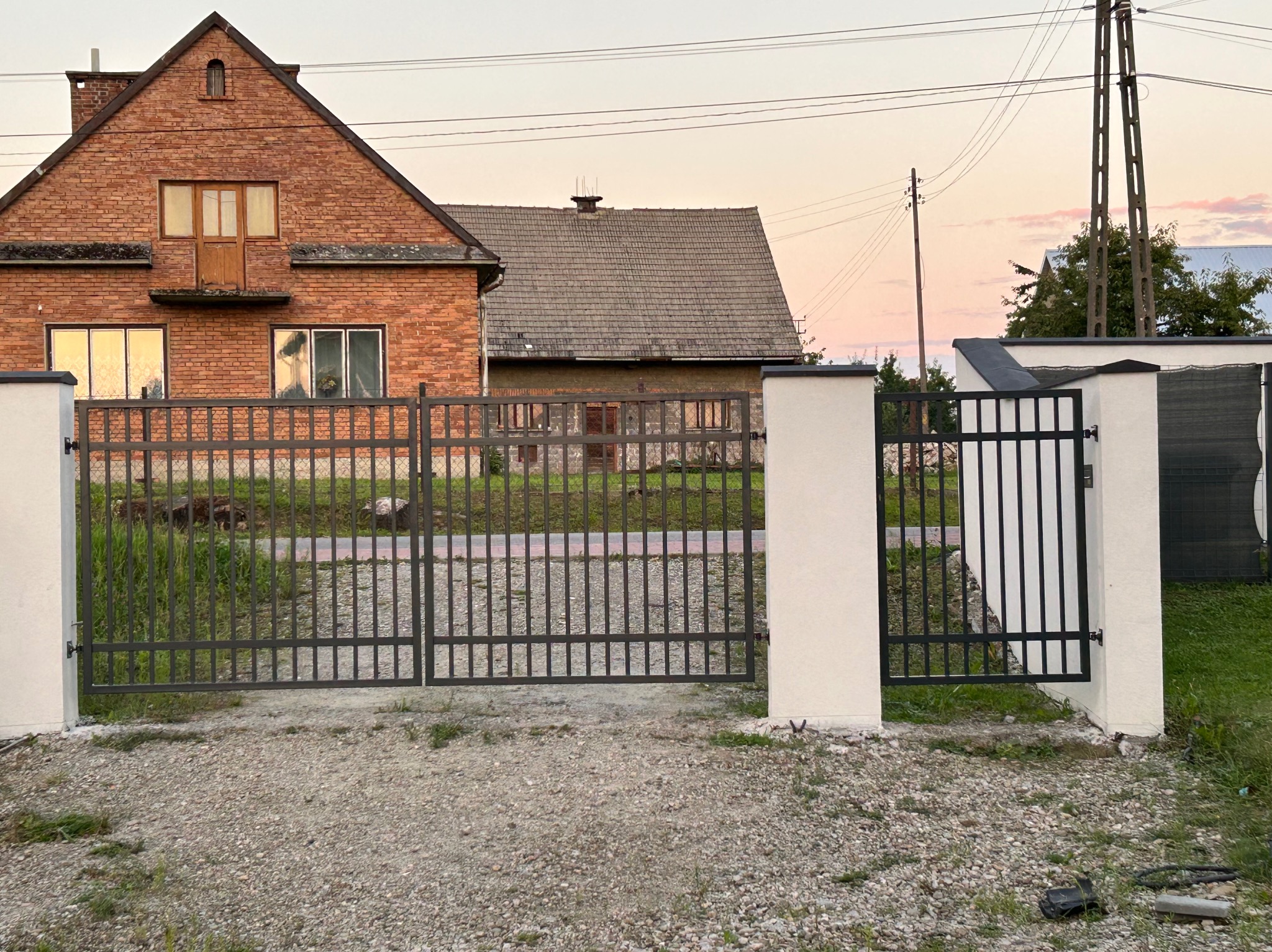 Nowoczesna, prosta brama wjazdowa z metalowymi prętami, osadzona pomiędzy białymi, murowanymi słupkami na tle ceglanego domu z drewnianymi elementami i żwirowego podjazdu.