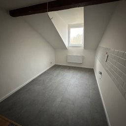 Zakład usług ogólno budowlanych Zalewski Sławomir - Poddasze po remoncie z szarą podłogą z płytek, świeżo pomalowanymi białymi ścianami, drewnianą belką stropową, białym grzejnikiem pod oknem i fragmentem ściany z białymi płytkami.