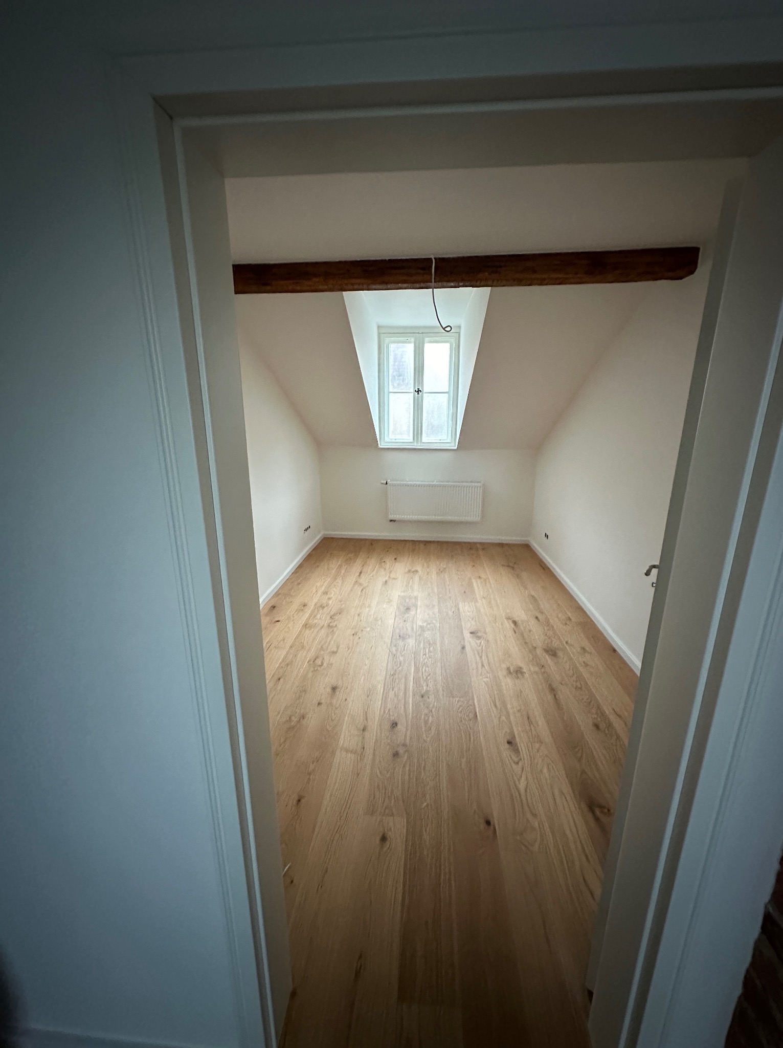 Poddasze z drewnianą belką stropową, jasnym parkietem i oknem w szczycie dachu, widziane zza framugi drzwi.